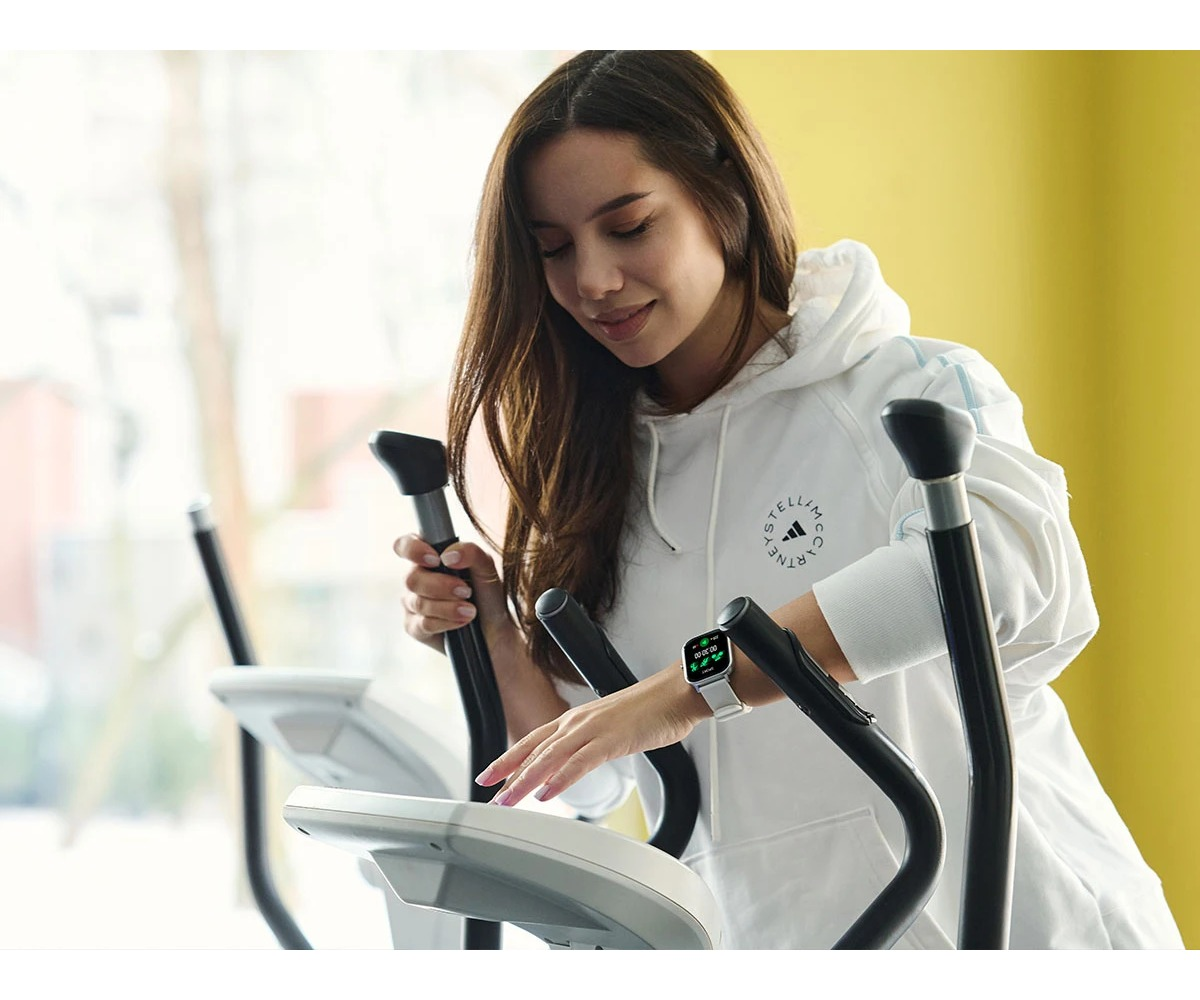 Una mujer con una sudadera blanca opera la consola de una elíptica en un gimnasio.