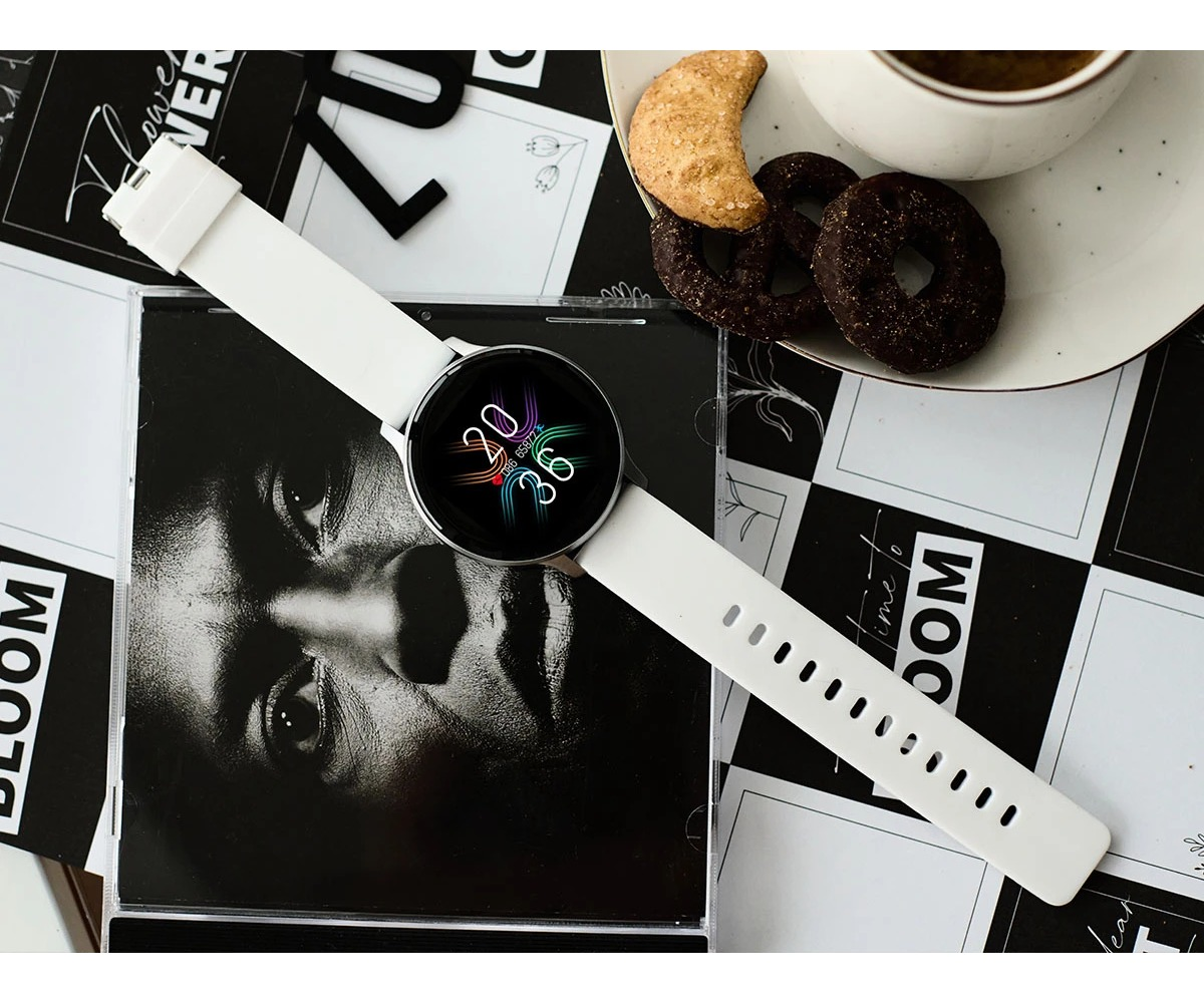 Un reloj inteligente blanco con números coloridos en la pantalla, un plato de galletas y una taza en una mesa en blanco y negro.