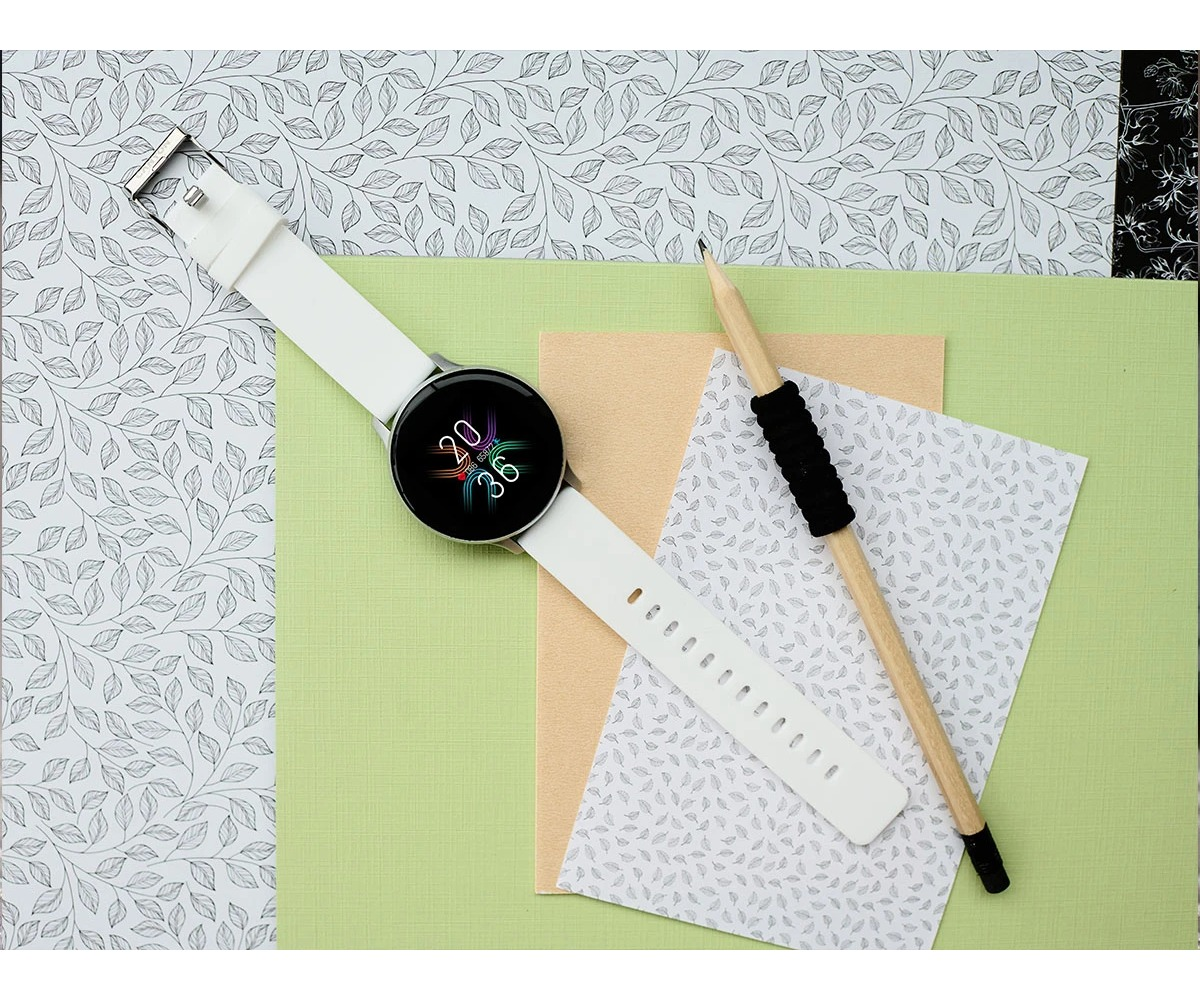 Un reloj inteligente blanco con pantalla colorida y un lápiz descansan sobre papeles estampados y verdes.