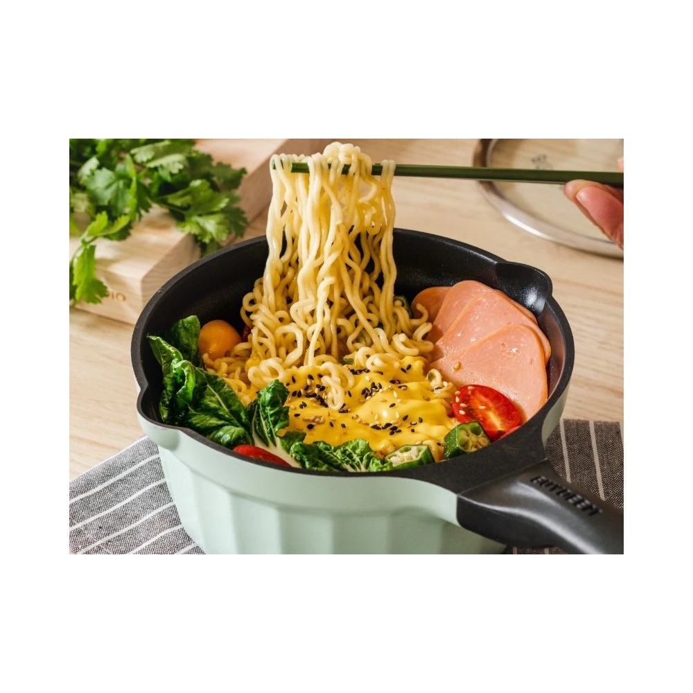 Makaron jest podnoszony pałeczkami z zielonej patelni z warzywami i szynką.