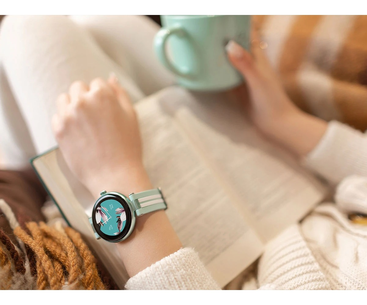 Osoba w białym ubraniu trzyma otwartą książkę, ma na sobie smartwatch i trzyma kubek.