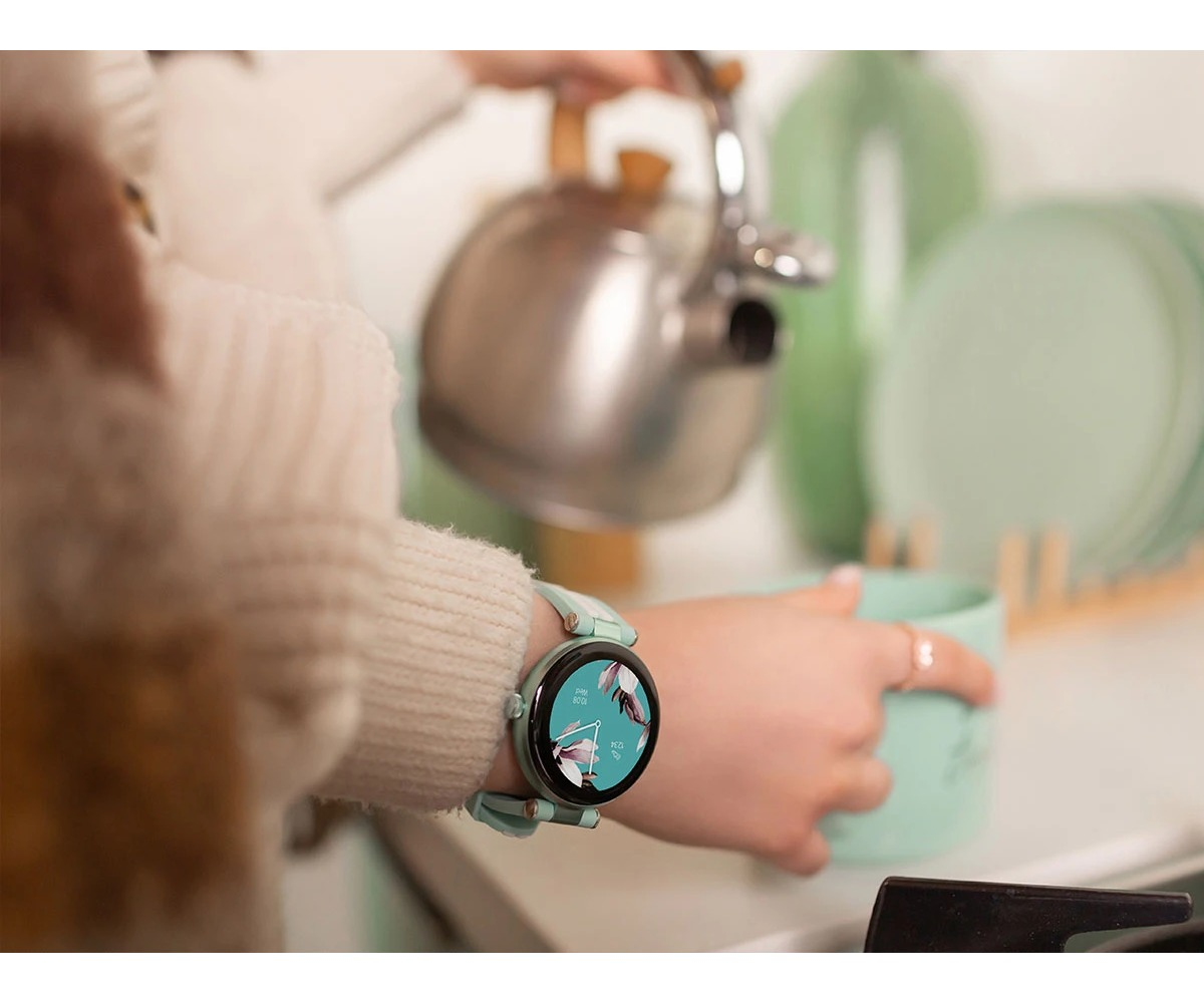 Osoba wlewa wodę ze srebrnego czajnika do zielonego kubka, mając na sobie jasny sweter i miętowy smartwatch.