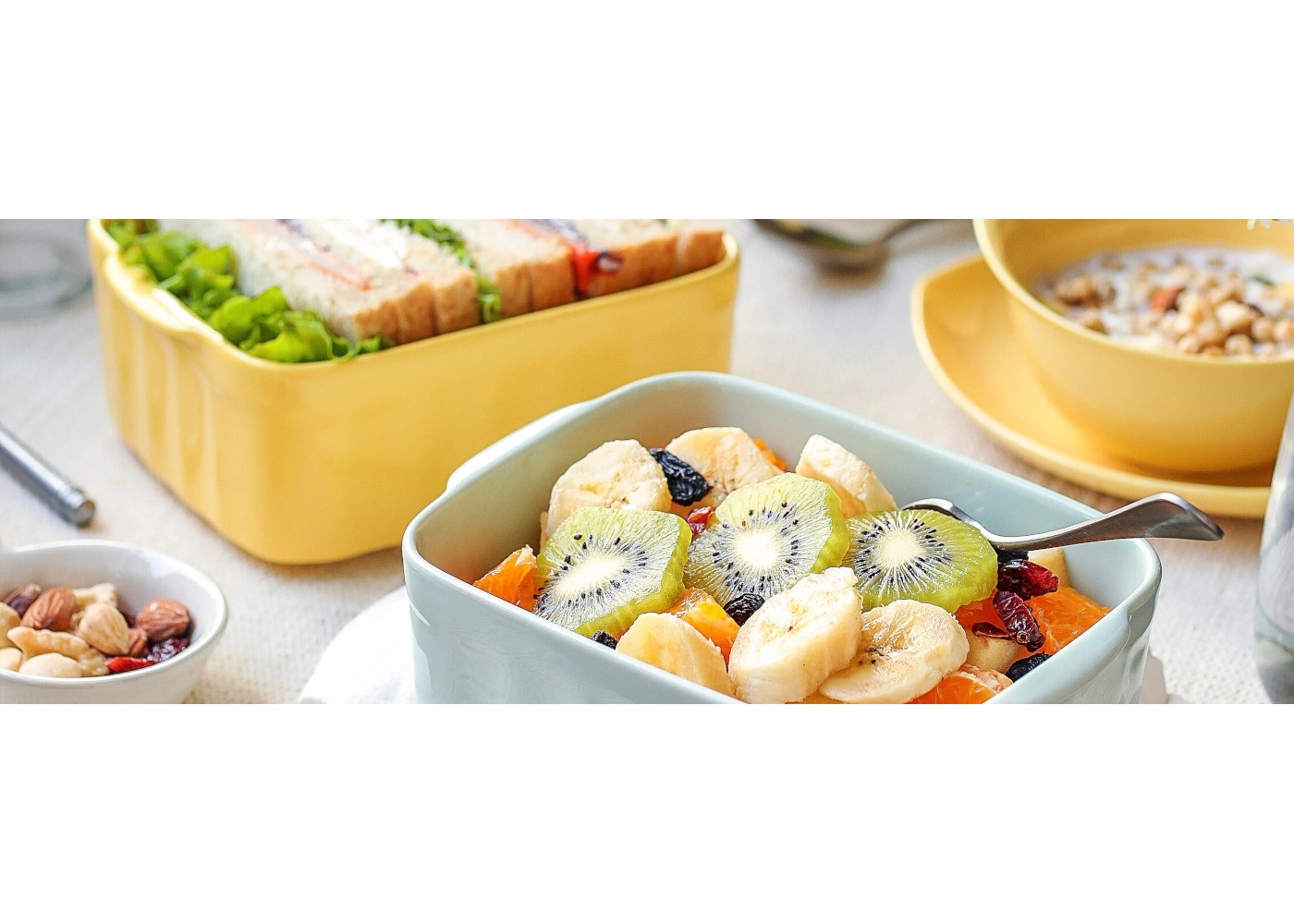 Miska pokrojonych bananów, kiwi, pomarańczy i rodzynek, w otoczeniu kanapek i płatków śniadaniowych.