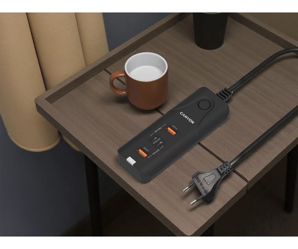 Czarna listwa zasilająca Canyon z portami USB leży na drewnianym stole obok kubka.