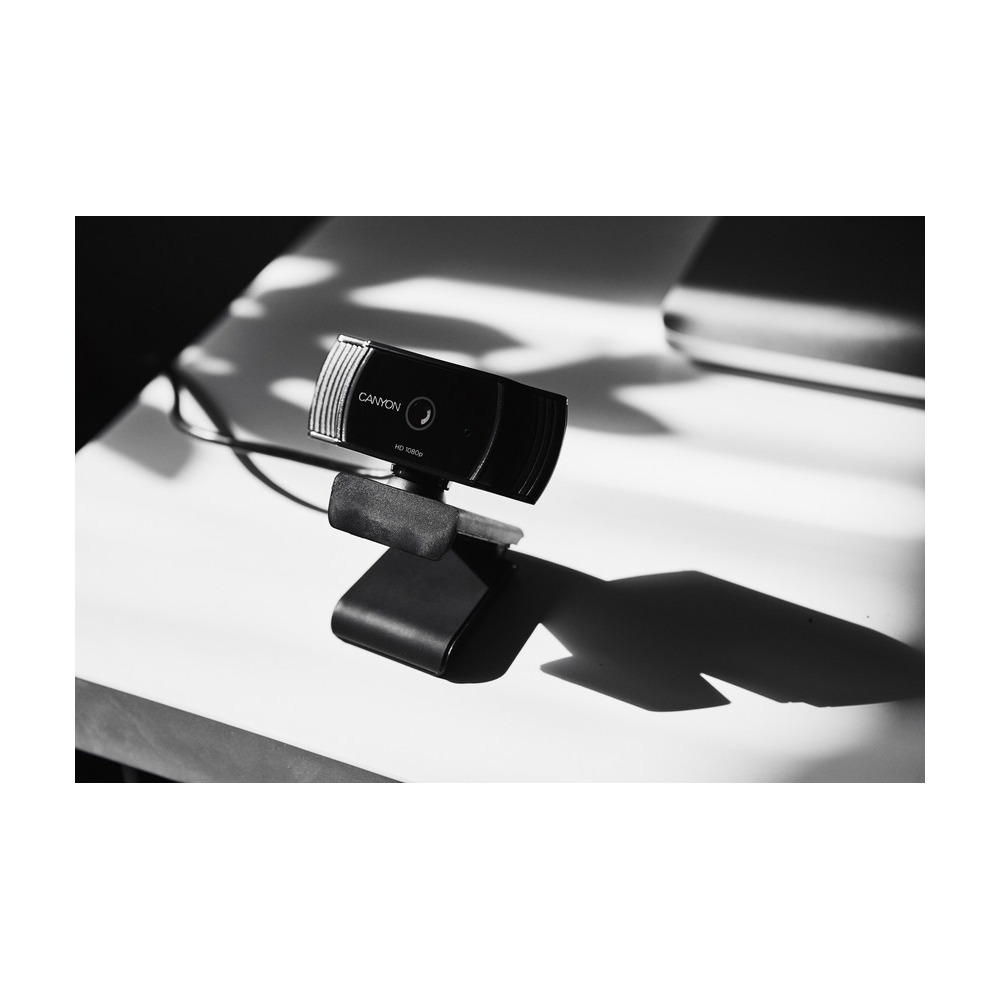 Foto en blanco y negro de una cámara web Canyon HD en un escritorio blanco con sombras marcadas.