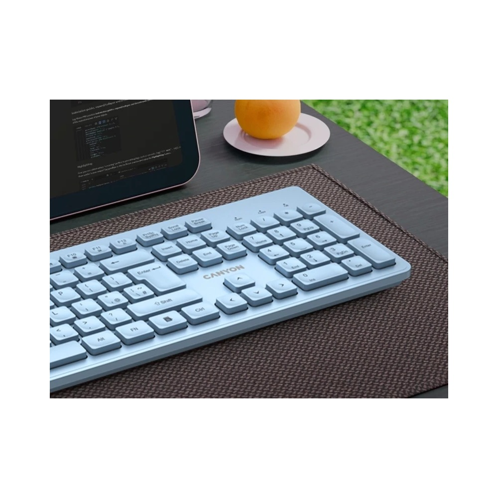 Jasnoniebieska klawiatura Canyon na pierwszym planie na brązowej podkładce, w tle tablet i pomarańcza.