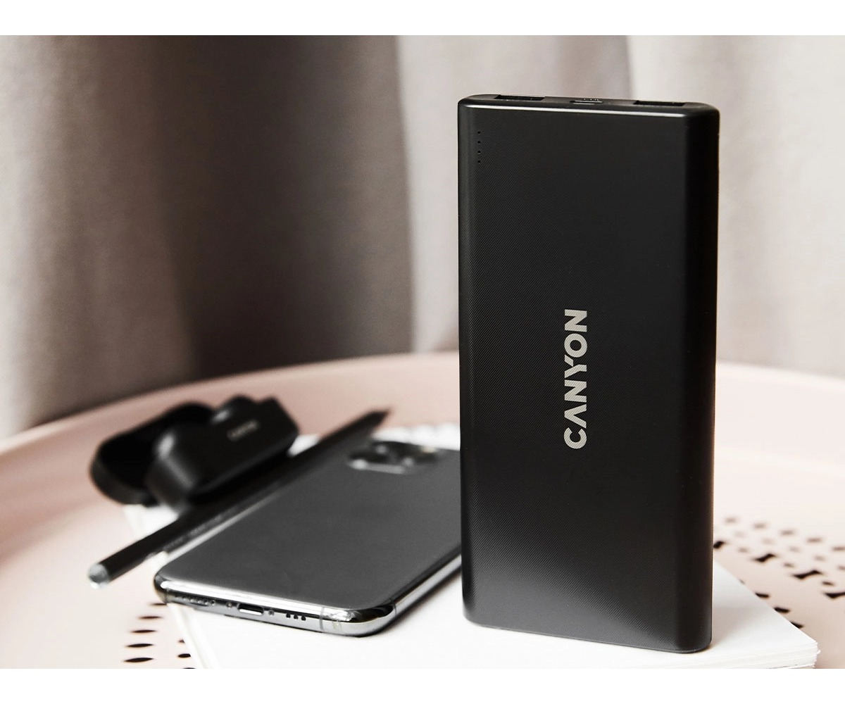 Czarny powerbank Canyon stoi na stole obok smartfona i słuchawek.