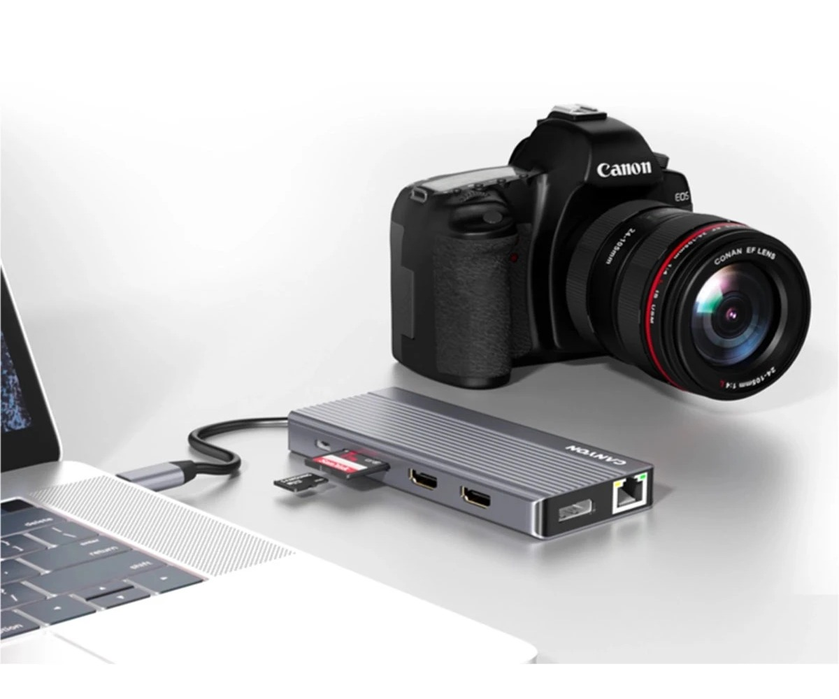 Szary hub USB-C podłączony do laptopa, z włożonymi kartami pamięci i czarnym aparatem Canon w tle.