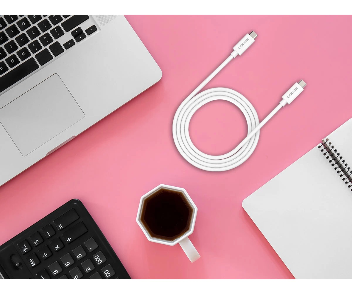 Biały kabel USB-C, laptop, kalkulator, notes i filiżanka kawy na różowym biurku.