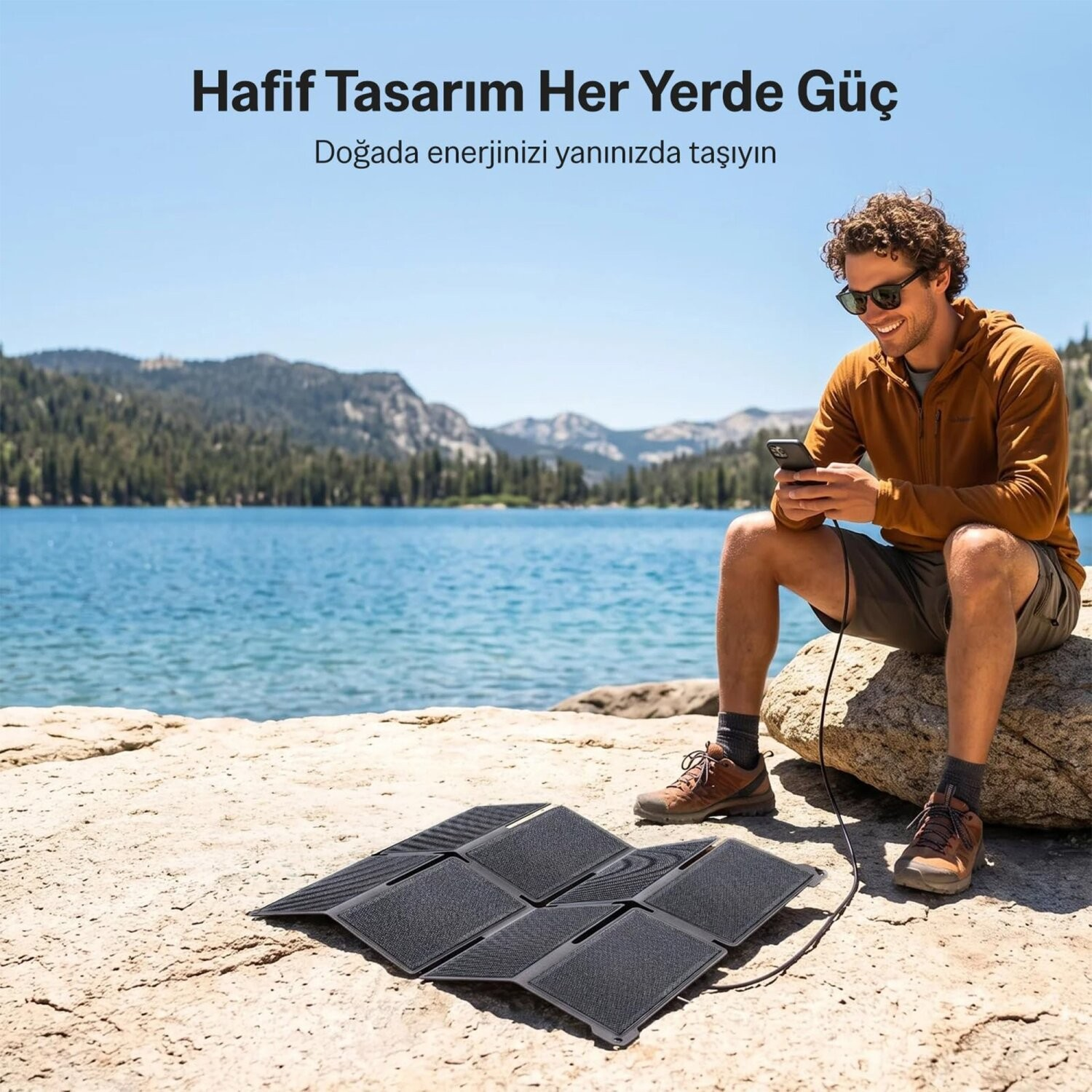 Bir adam göl kenarındaki kayada oturmuş, taşınabilir güneş paneliyle telefonunu şarj ediyor.