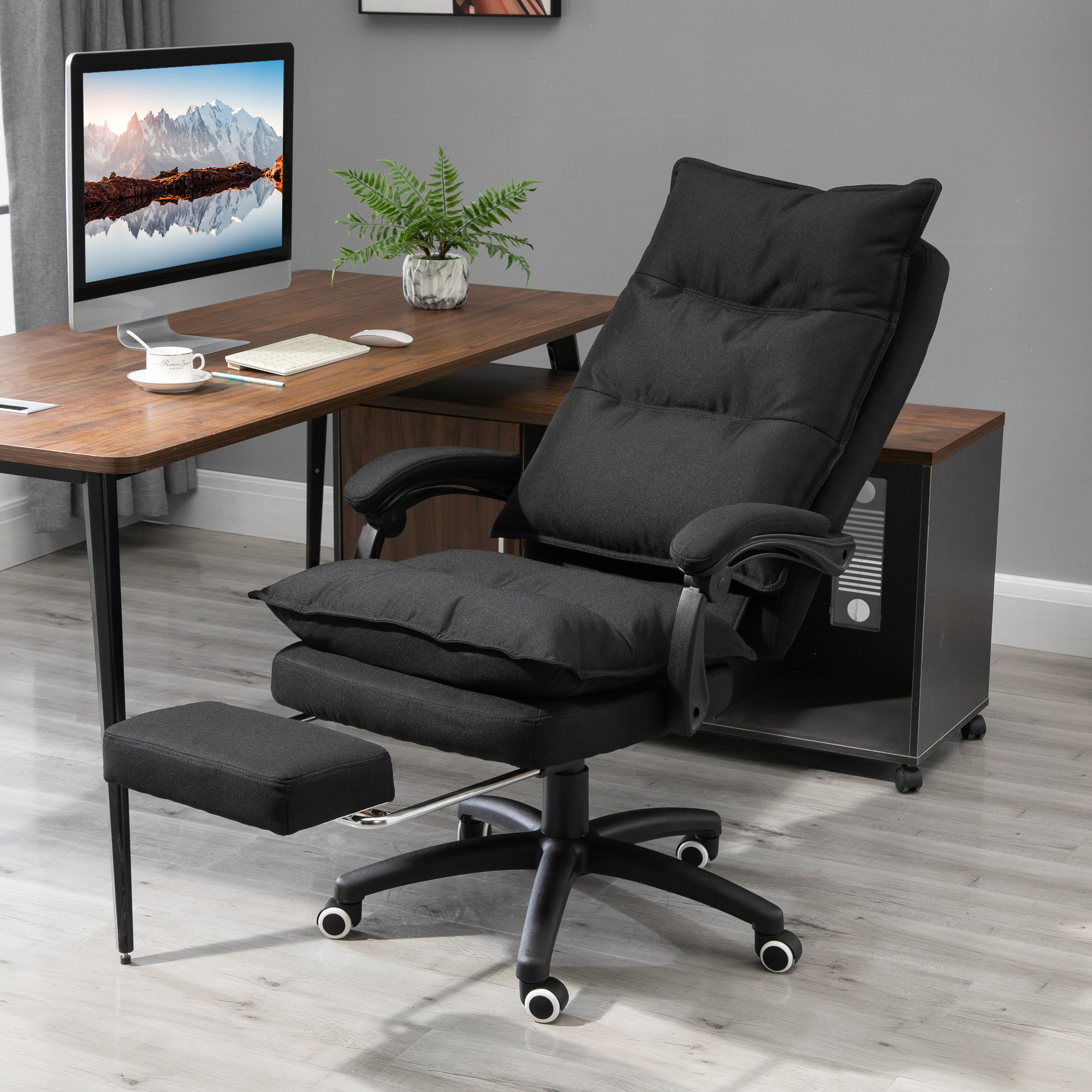 Una sedia da ufficio ergonomica nera con poggiapiedi si trova a una scrivania in legno con PC.