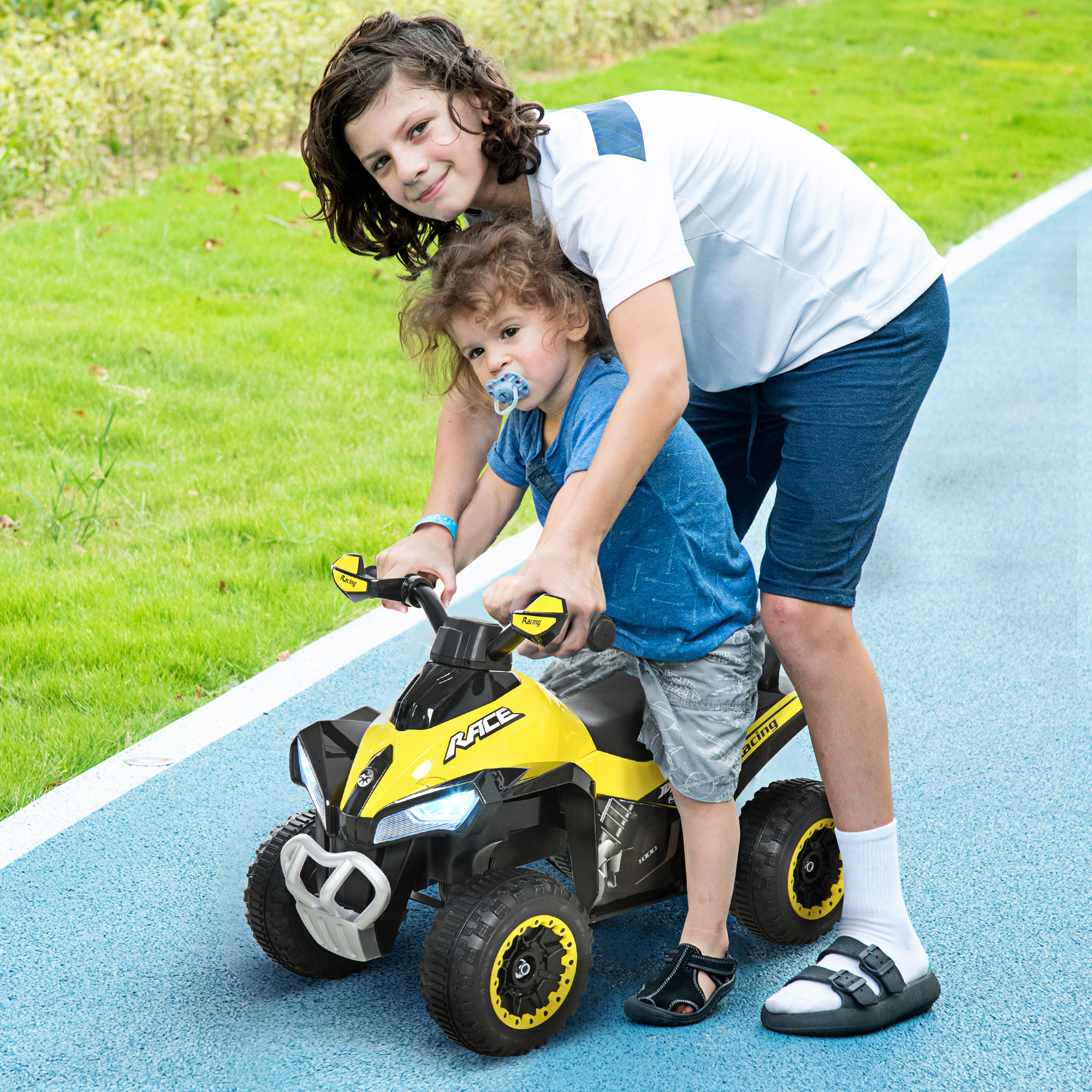 Un bambino più grande aiuta un piccolo con il ciuccio a guidare un quad giallo e nero.