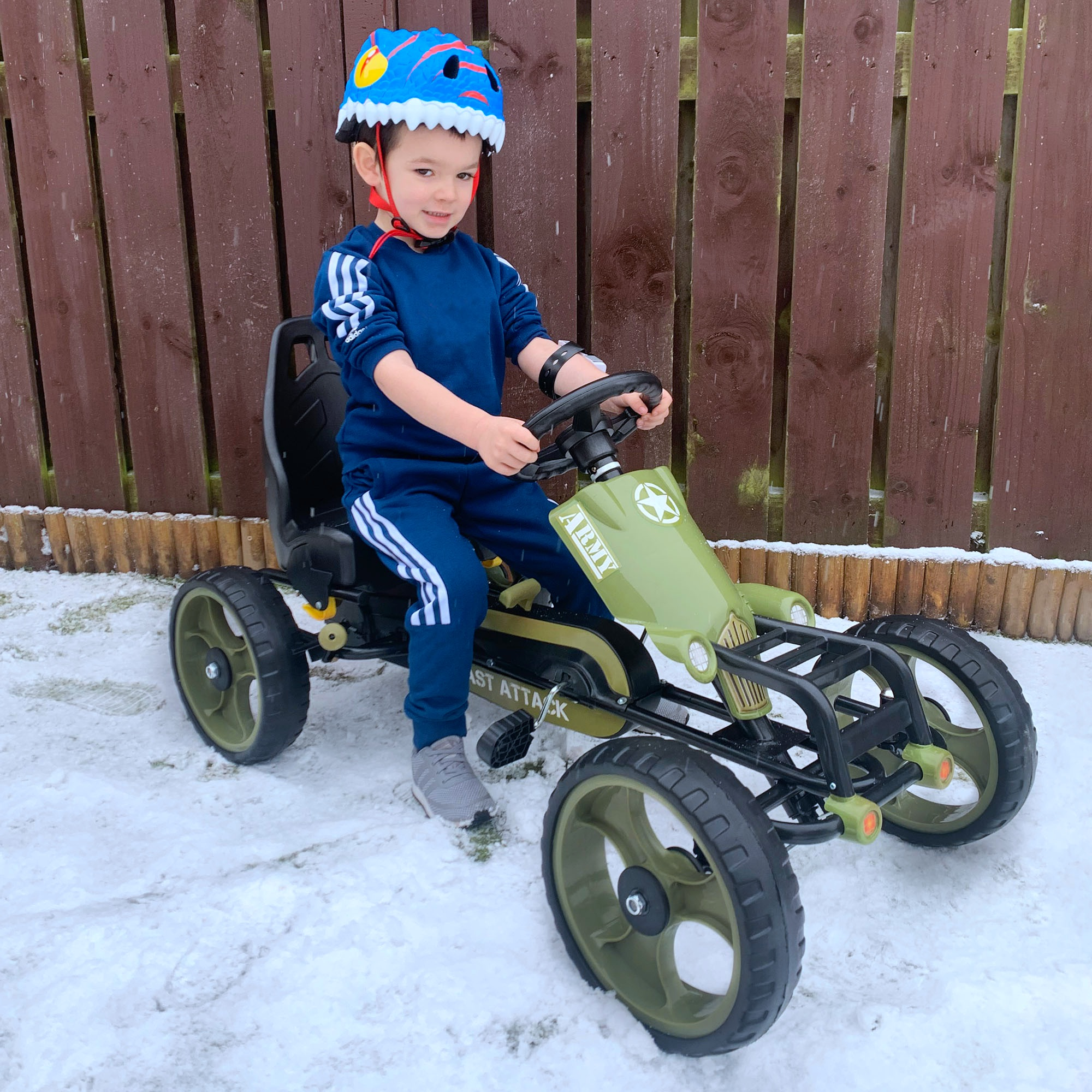Un bambino in tuta blu e casco da dinosauro guida un go-kart verde sulla neve davanti a un recinto in legno.