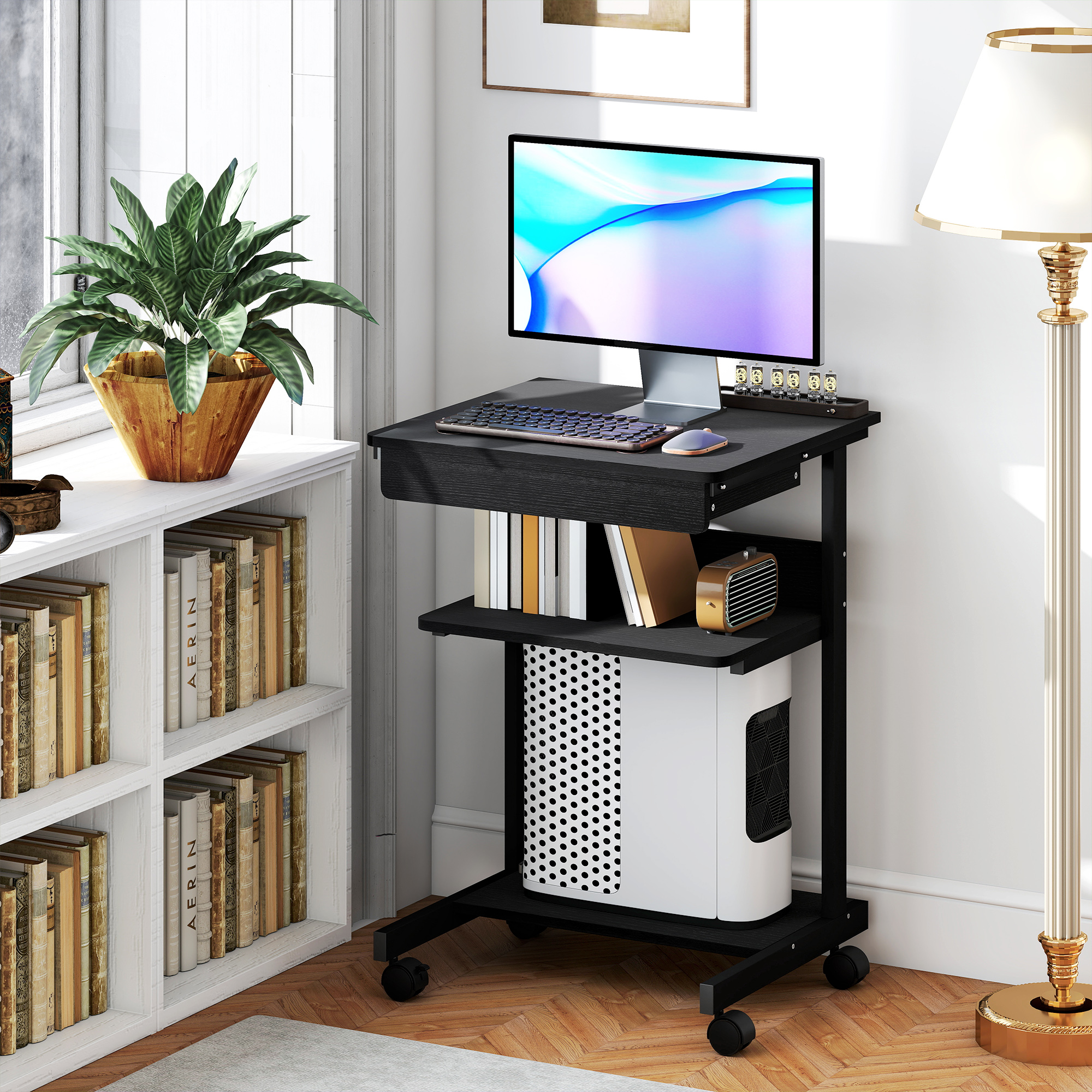 Un carrello per computer nero con ruote, monitor, tastiera e torre PC, accanto a una libreria bianca e una lampada.