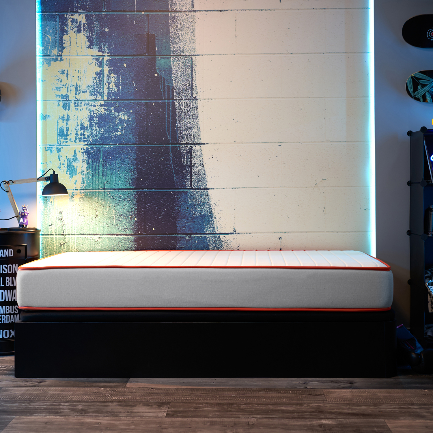 Una cama con colchón blanco y borde naranja se encuentra contra una pared de ladrillo pintada de azul y blanco con luz de neón.