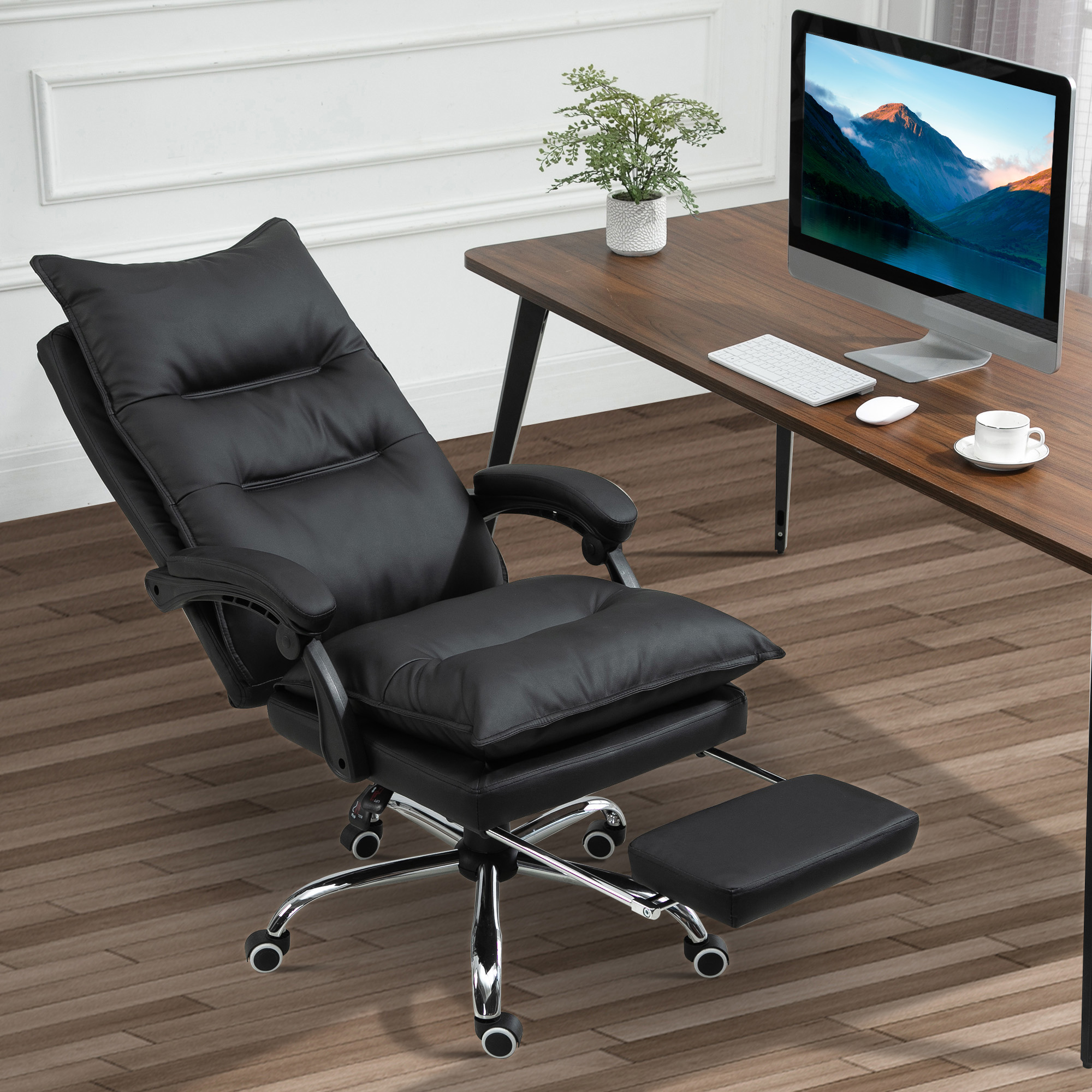 Una sedia da ufficio in pelle nera con poggiapiedi è davanti a una scrivania in legno con computer, pianta e tazza.