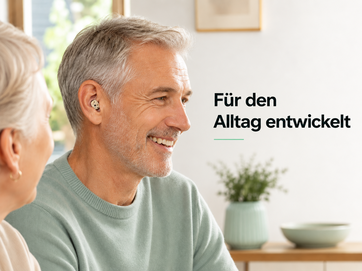 Älterer Mann lächelt, trägt ein kleines Hörgerät im Ohr. Im Vordergrund sitzt eine Frau, im Hintergrund ein Tisch mit Pflanze.