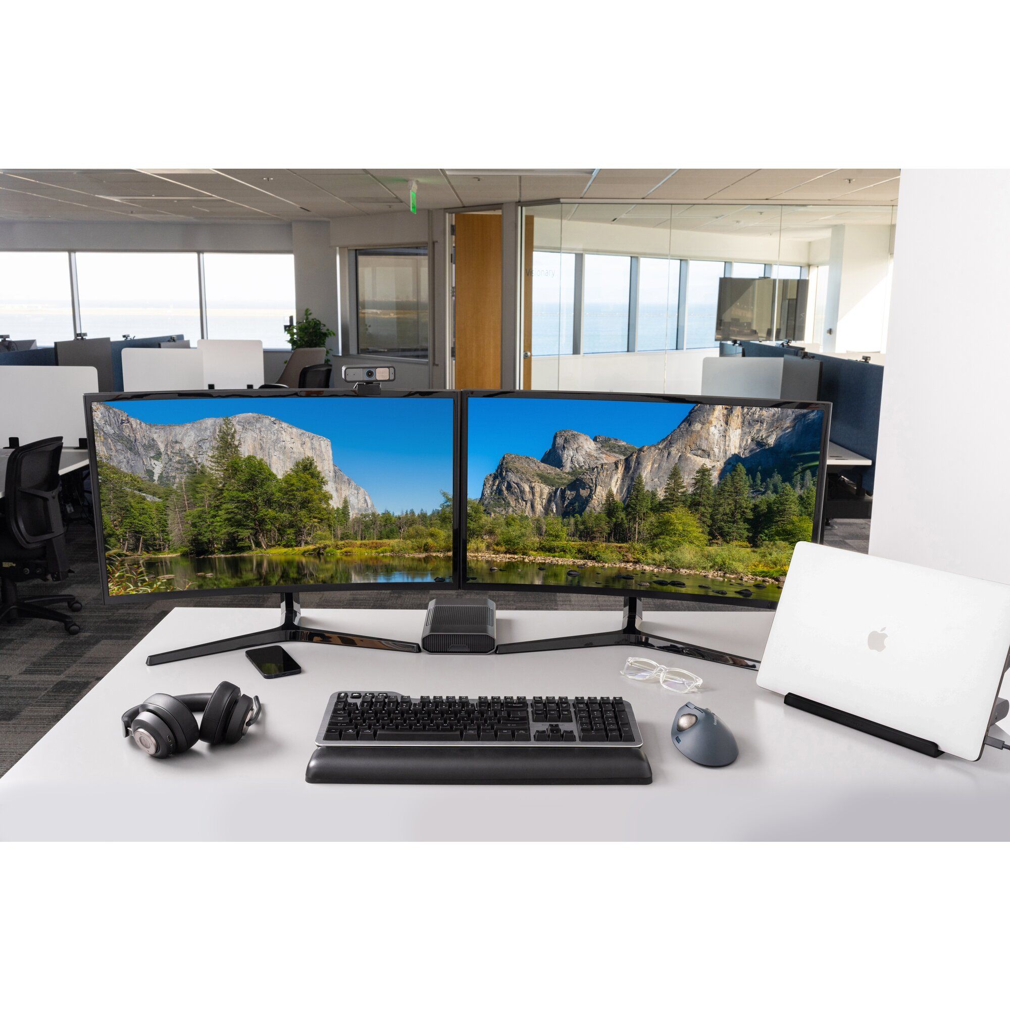 Office desk with dual monitors, keyboard, mouse, headphones, and laptop in a bright workspace.