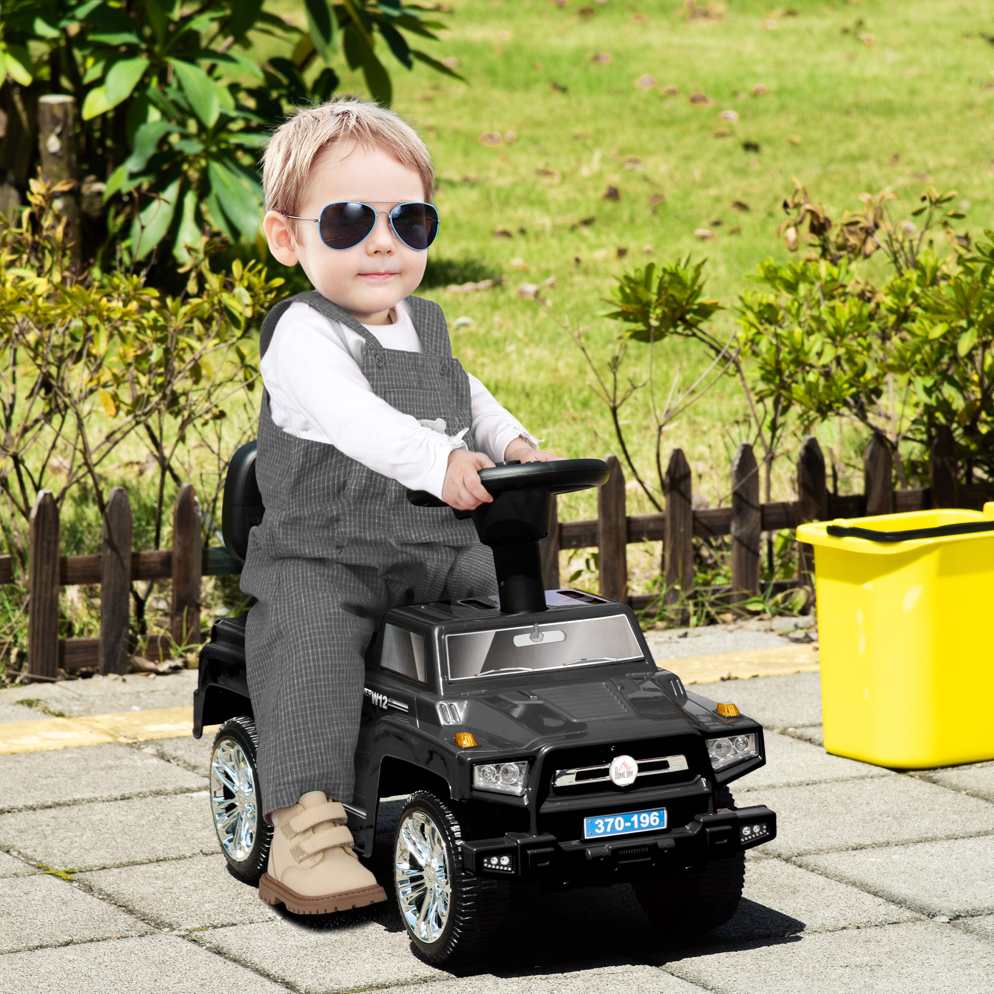 Un bambino con occhiali da sole siede su un SUV giocattolo nero all'aperto, vicino a un bidone giallo.