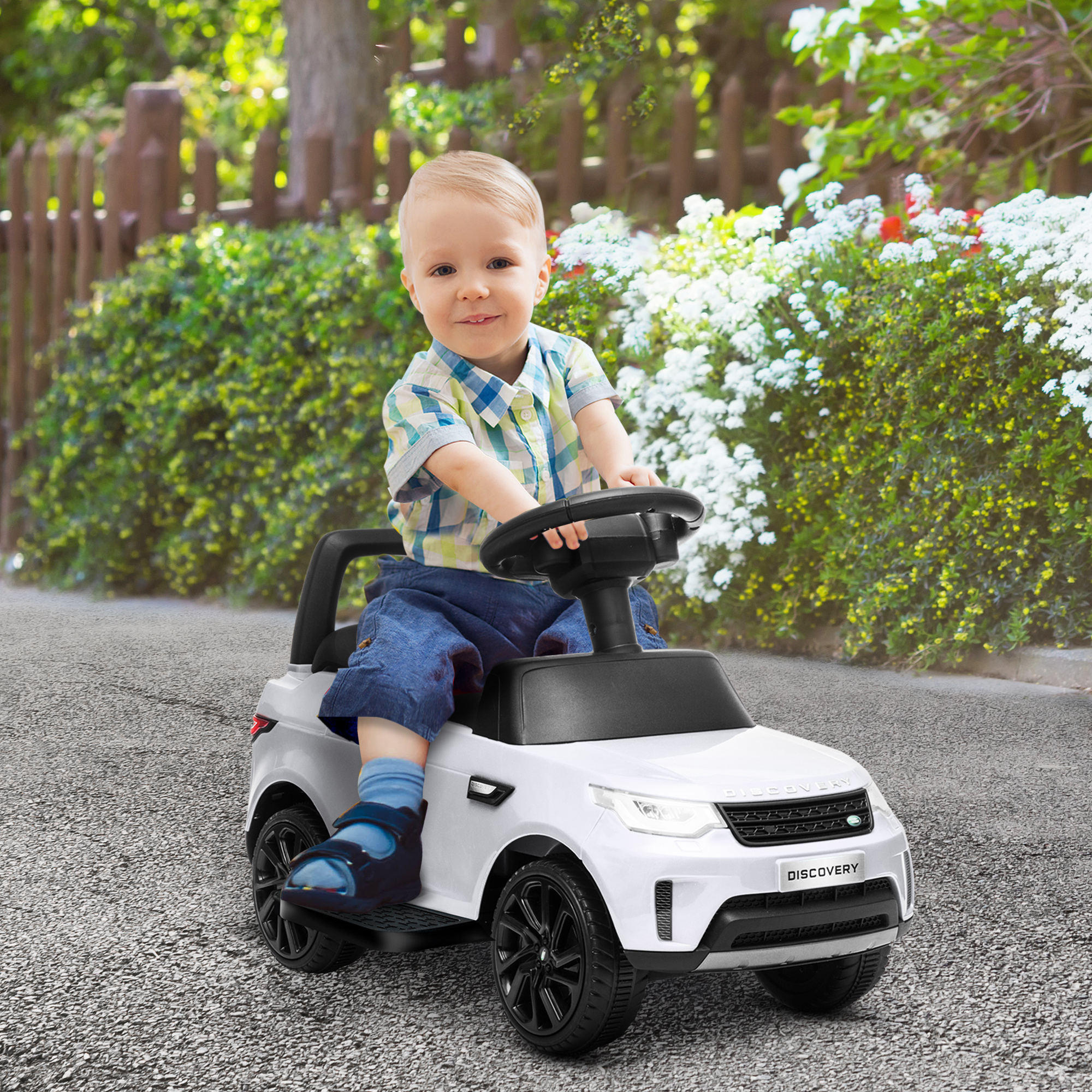 Un bambino sorride mentre guida un'auto giocattolo bianca all'aperto davanti a un giardino.