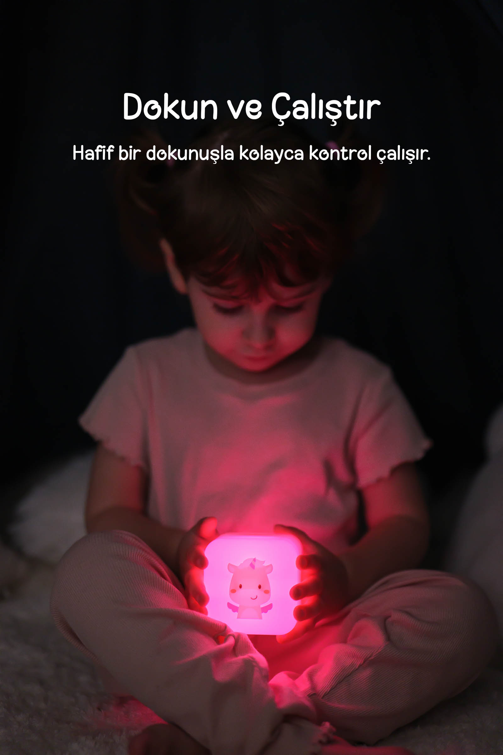 Karanlıkta oturan küçük bir kız, üzerinde hayvan motifi olan pembe gece lambasını tutuyor.