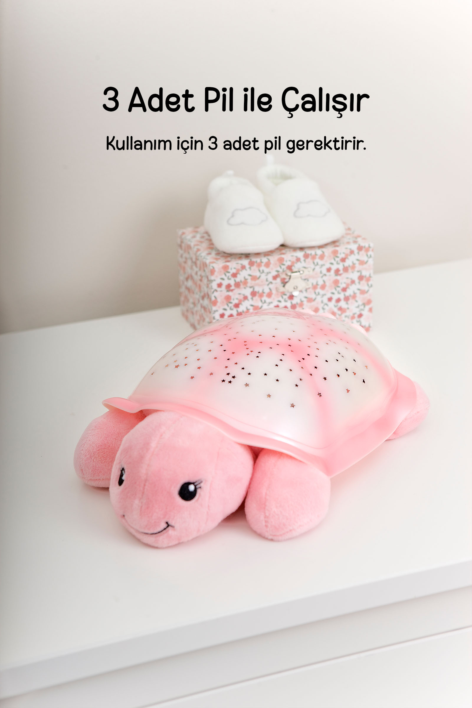 Yıldız kesikli pembe peluş kaplumbağa gece lambası beyaz zemin üstünde, arkada çiçekli kutu ve bebek ayakkabıları ile duruyor.