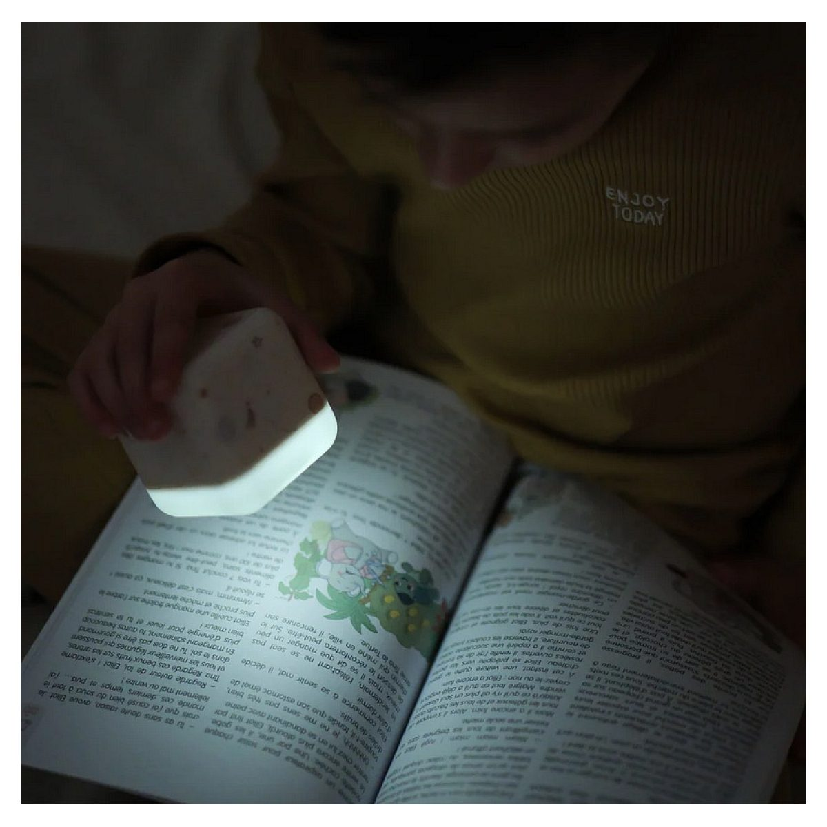Sarı kazaklı bir çocuk karanlıkta kitap okuyor, sayfaları aydınlatmak için küçük kare bir gece lambası kullanıyor.