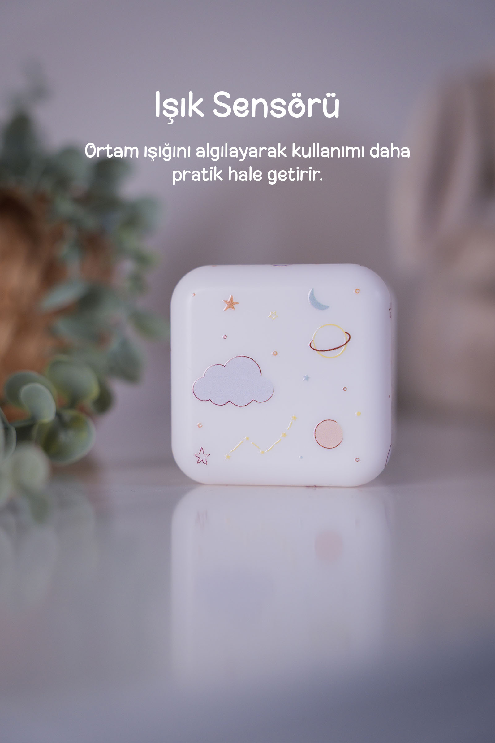 Bulut ve yıldız desenli beyaz bir gece lambası, bulanık bitkilerle çevrili bir masanın üzerinde duruyor.