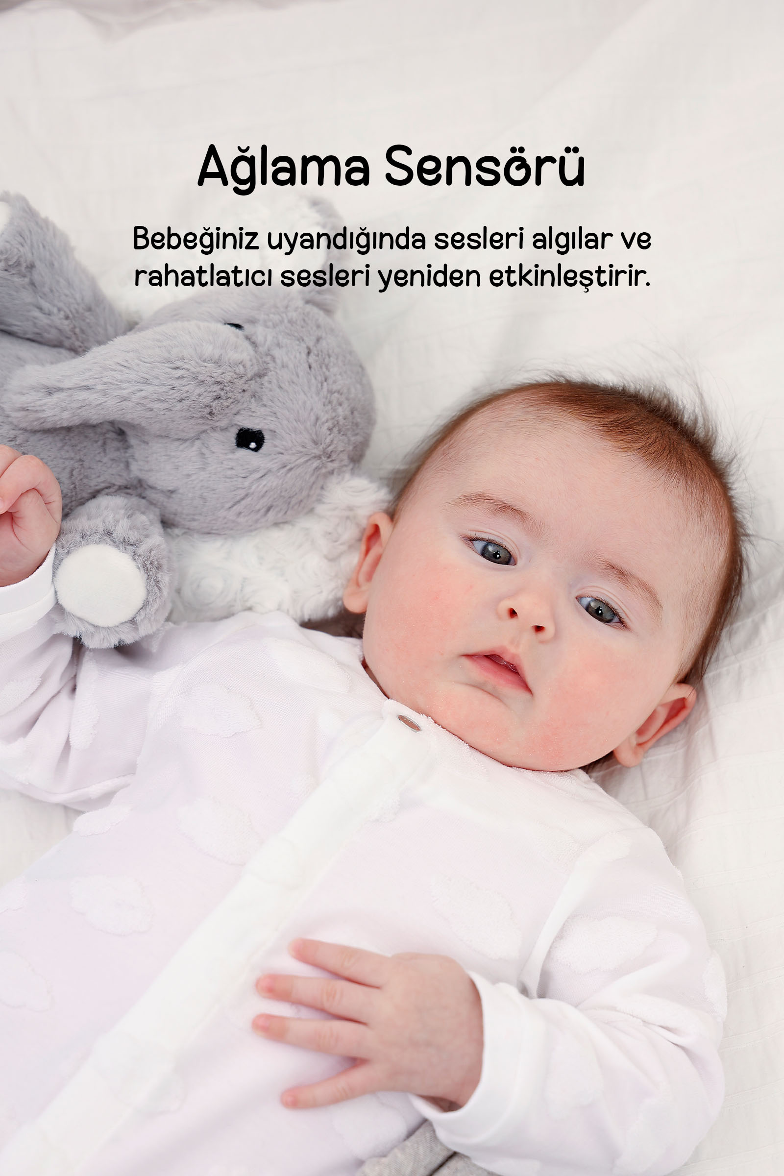 Beyaz pijamalı bebek, beyaz yatakta gri peluş tavşanın yanında yatıyor.