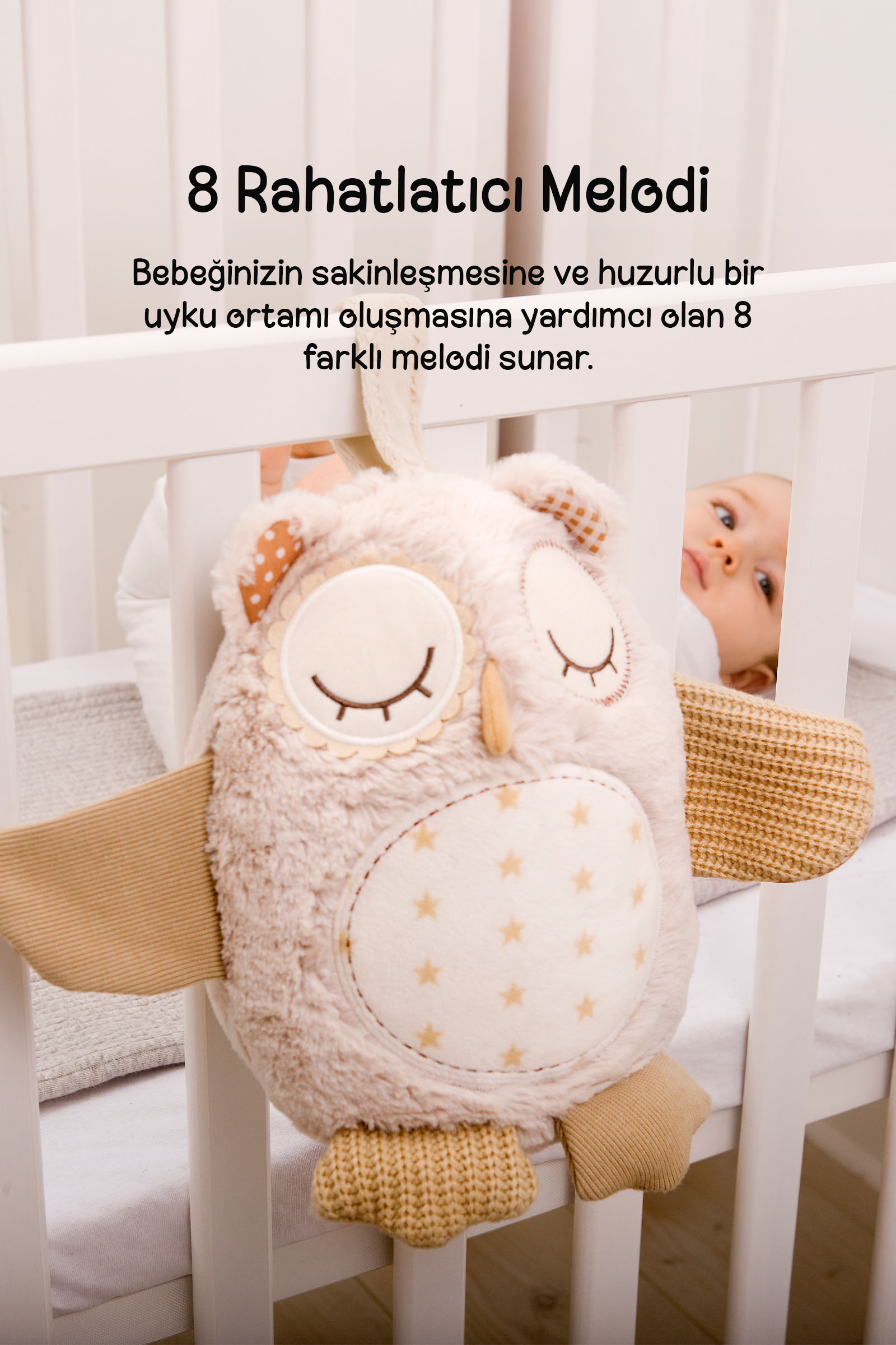 Beyaz bir bebek yatağında asılı peluş baykuş ve arkasında bakan bir bebek.