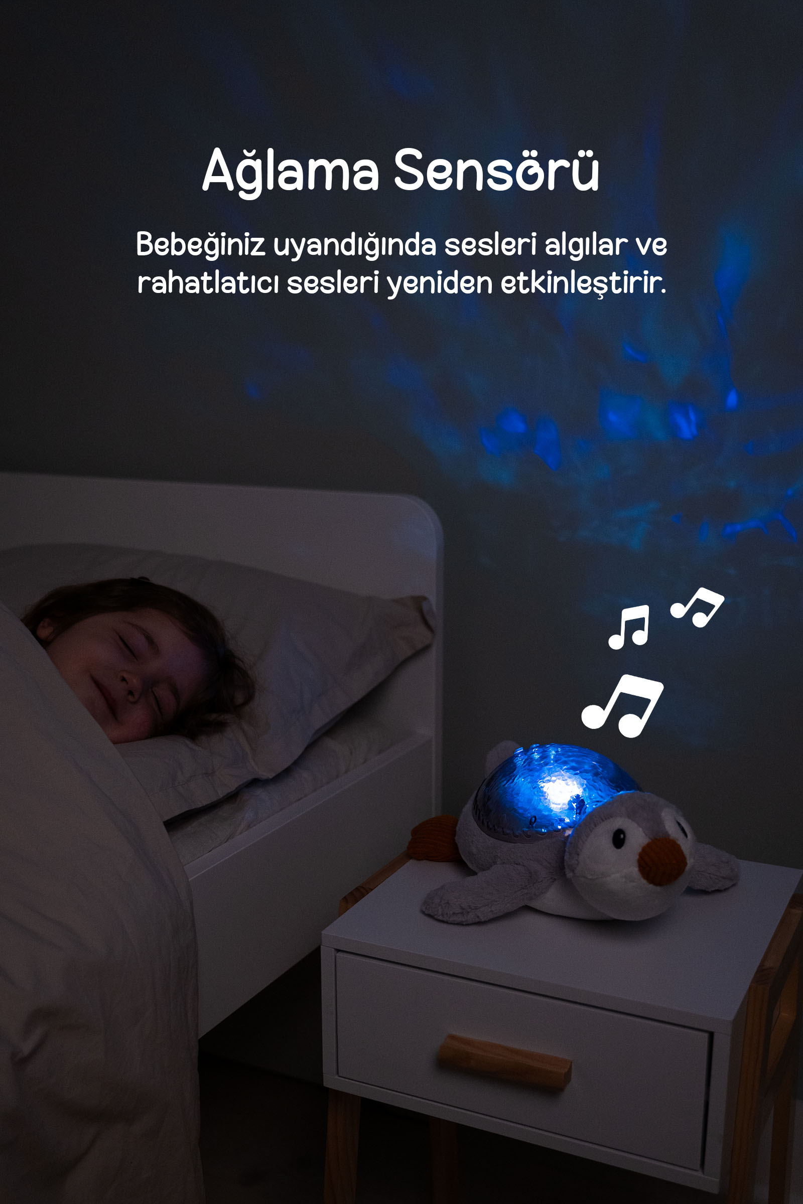 Mavi projeksiyonlu ve nota ikonlu penguen gece lambası komodinde dururken bir çocuk yatakta uyuyor.