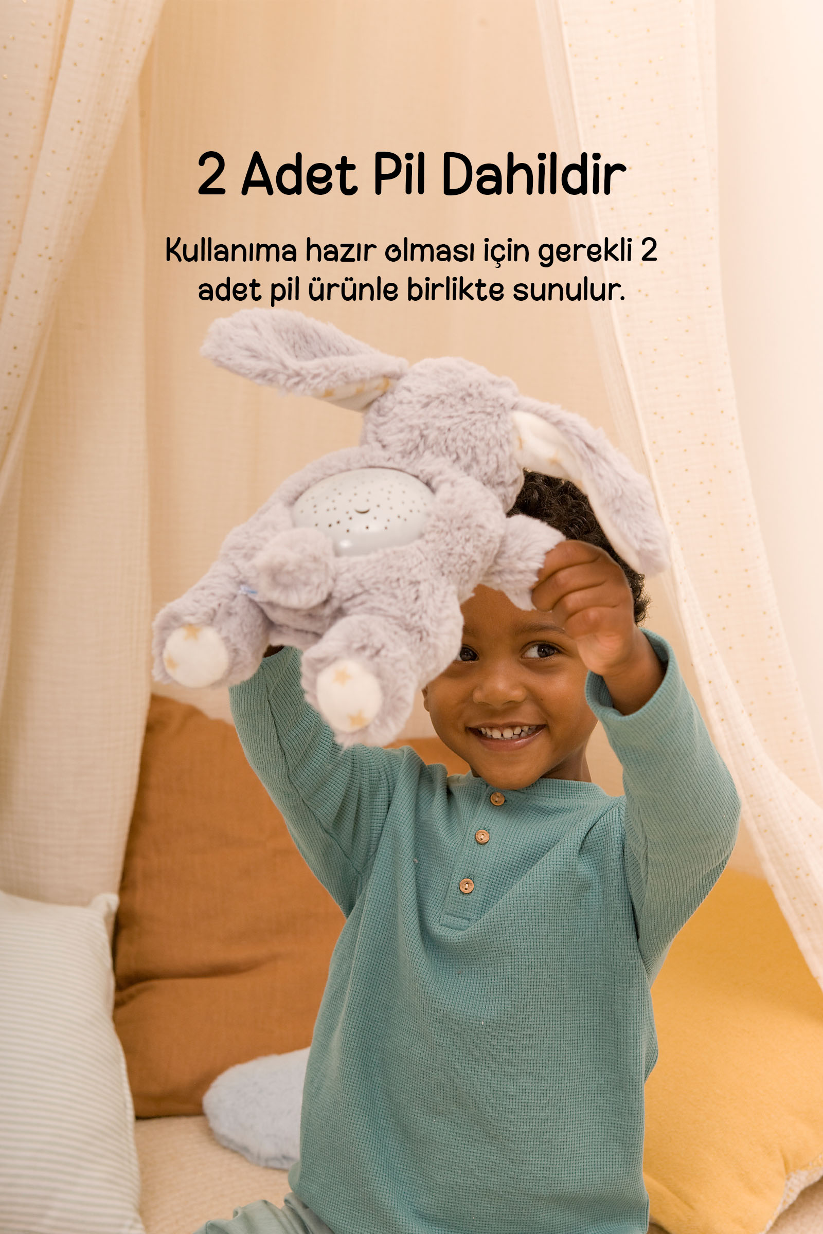 Nane yeşili kazaklı gülen bir çocuk, arka planda cibinlik ile gri peluş tavşan oyuncağı kaldırıyor.