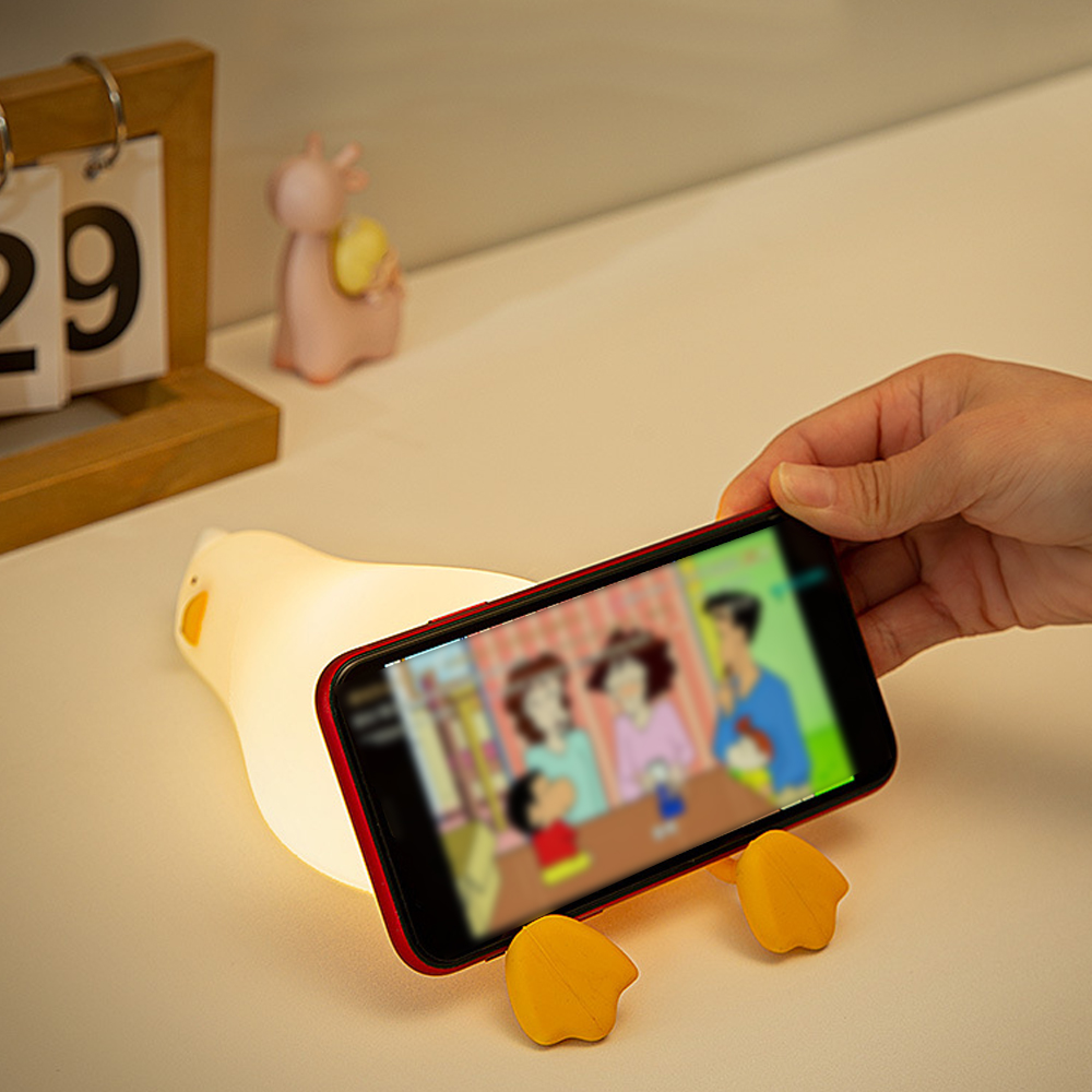 Eine Hand hält ein Smartphone in einem Enten-Halter auf einem Tisch mit einem kleinen Kalender.