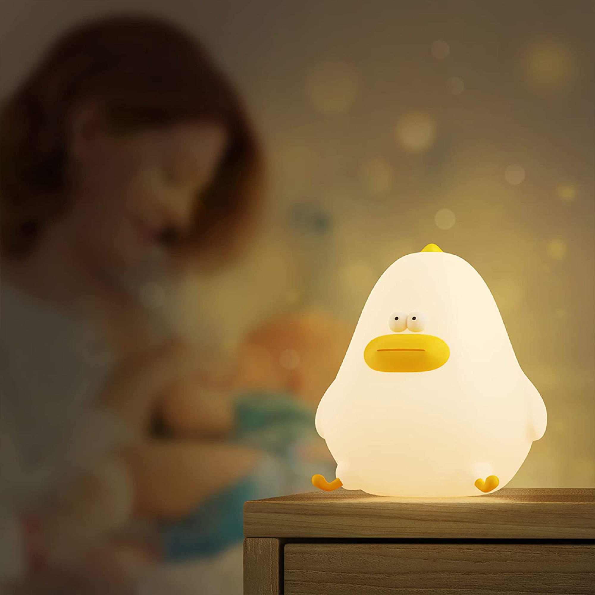 Ein leuchtendes entenförmiges Nachtlicht steht auf einem Holztisch, im Hintergrund eine verschwommene Mutter mit Baby.