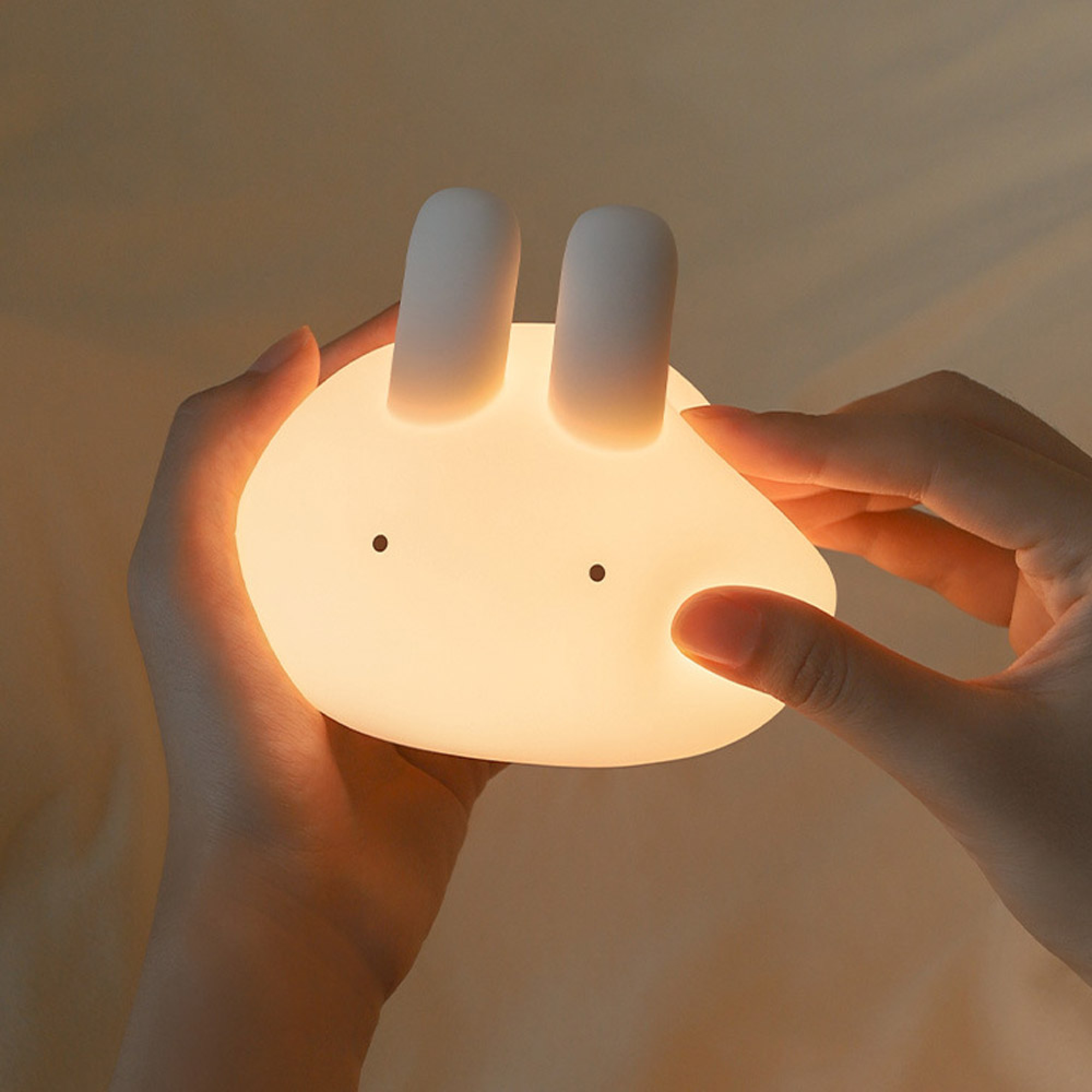 Hände halten eine weiche, leuchtende Hasenlampe mit zwei Ohren und schwarzen Augen.