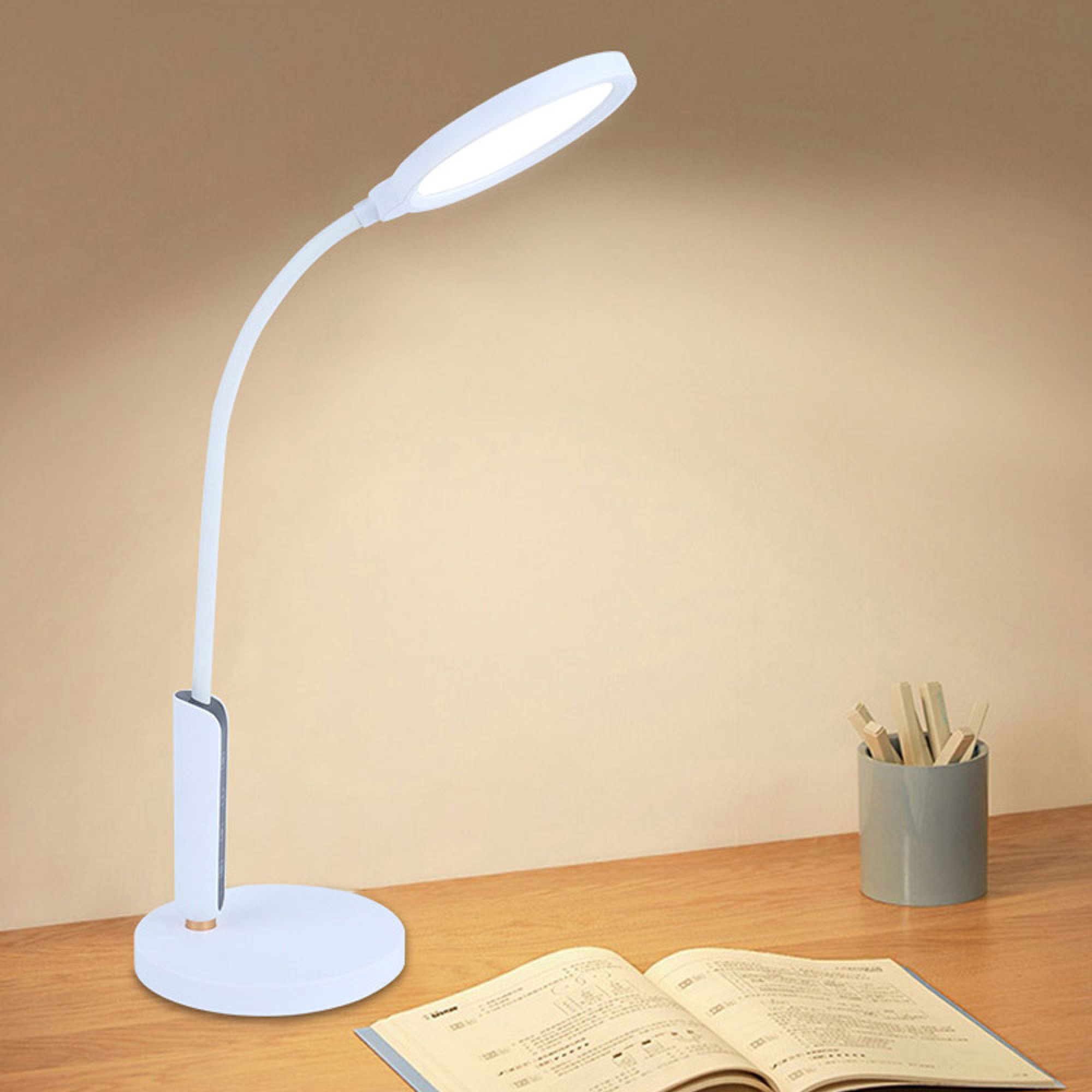 Eine weiße LED-Schreibtischlampe leuchtet auf einem Holztisch mit einem Buch und einem Stiftehalter im Hintergrund.