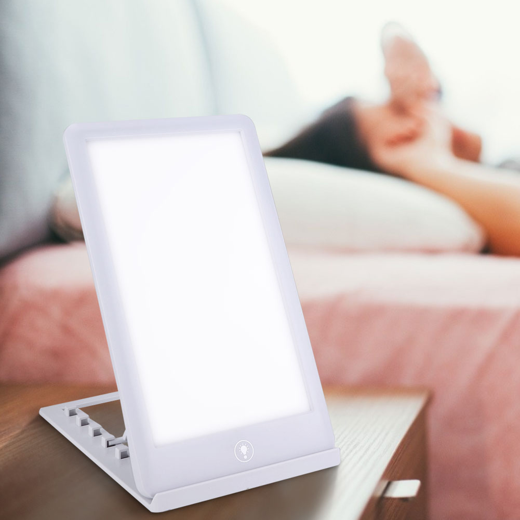 Eine weiße Tageslichtlampe steht auf einem Nachttisch, im Hintergrund ruht eine Person im Bett.