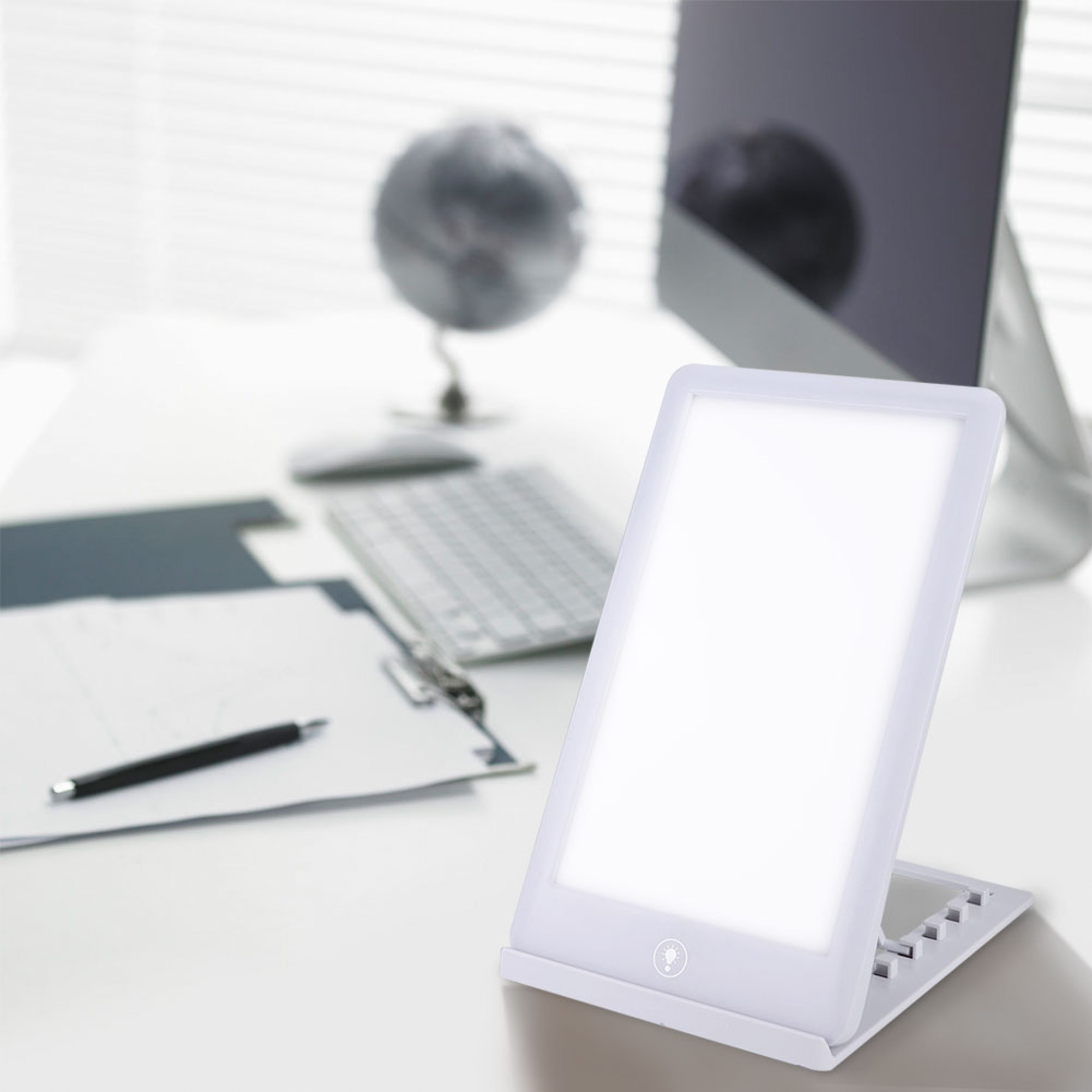 Eine weiße Tageslichtlampe auf einem Schreibtisch mit Monitor, Tastatur und Papieren im Hintergrund.