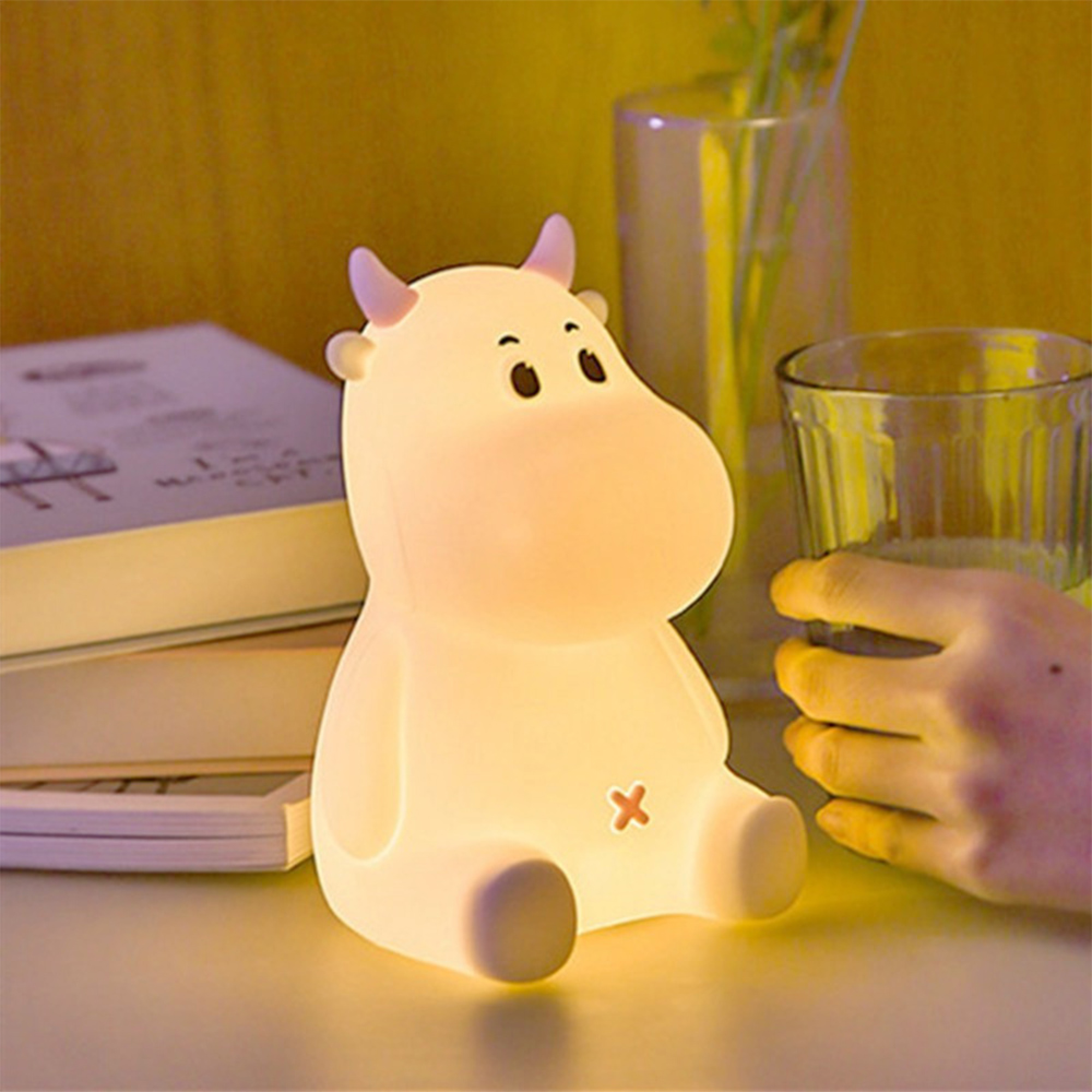 Eine niedliche, leuchtende Kuh-Lampe sitzt auf einem Tisch neben Büchern und einer Hand, die ein Glas hält.