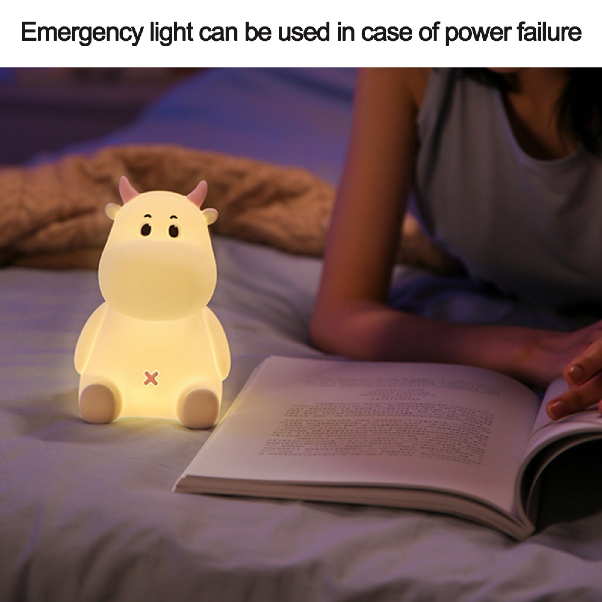 Eine süße, leuchtende Kuh-Nachtlampe steht auf dem Bett neben einem offenen Buch, während eine Person liest.