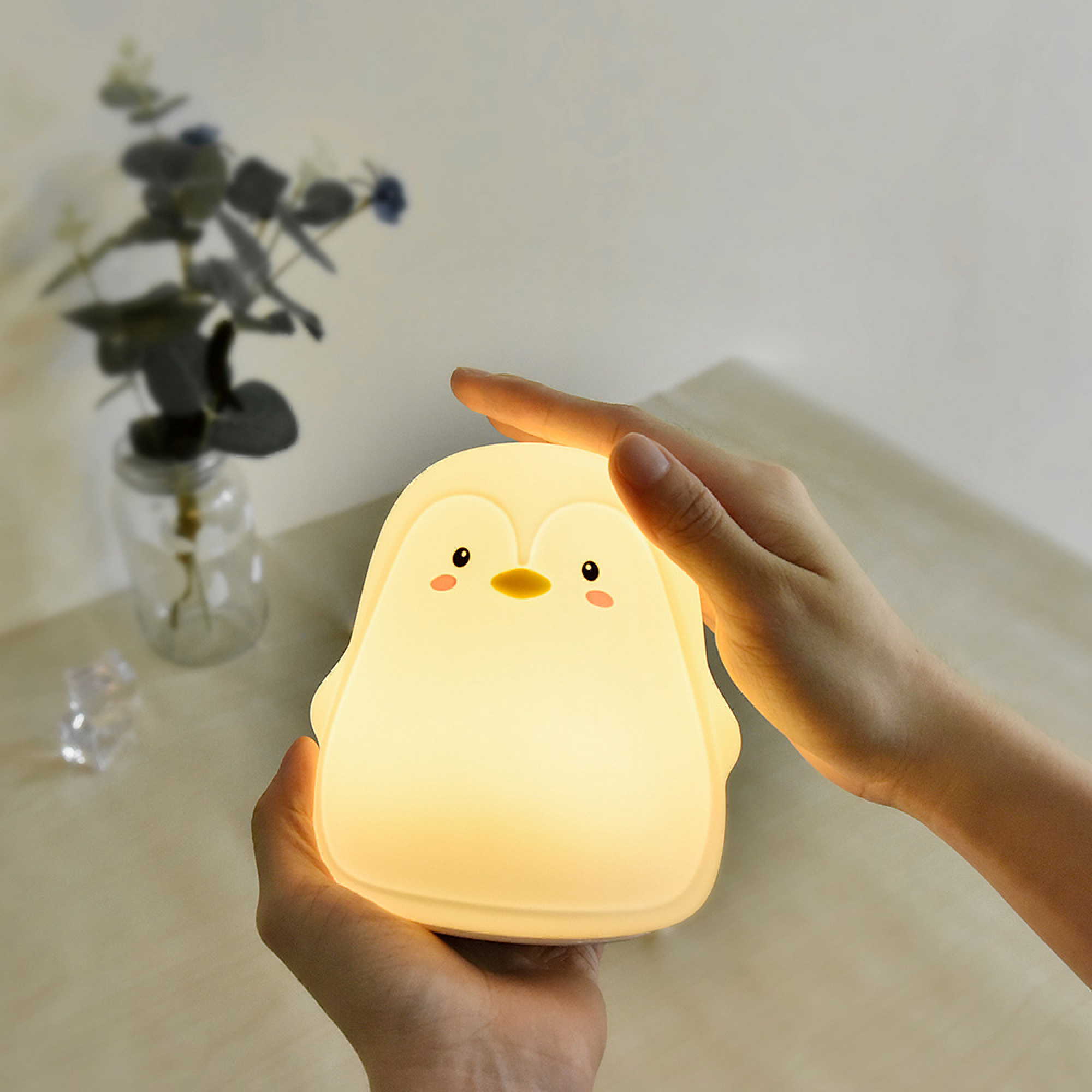 Eine Hand hält eine niedliche, leuchtende Pinguin-Nachtlampe auf einem Holztisch, im Hintergrund eine Vase.