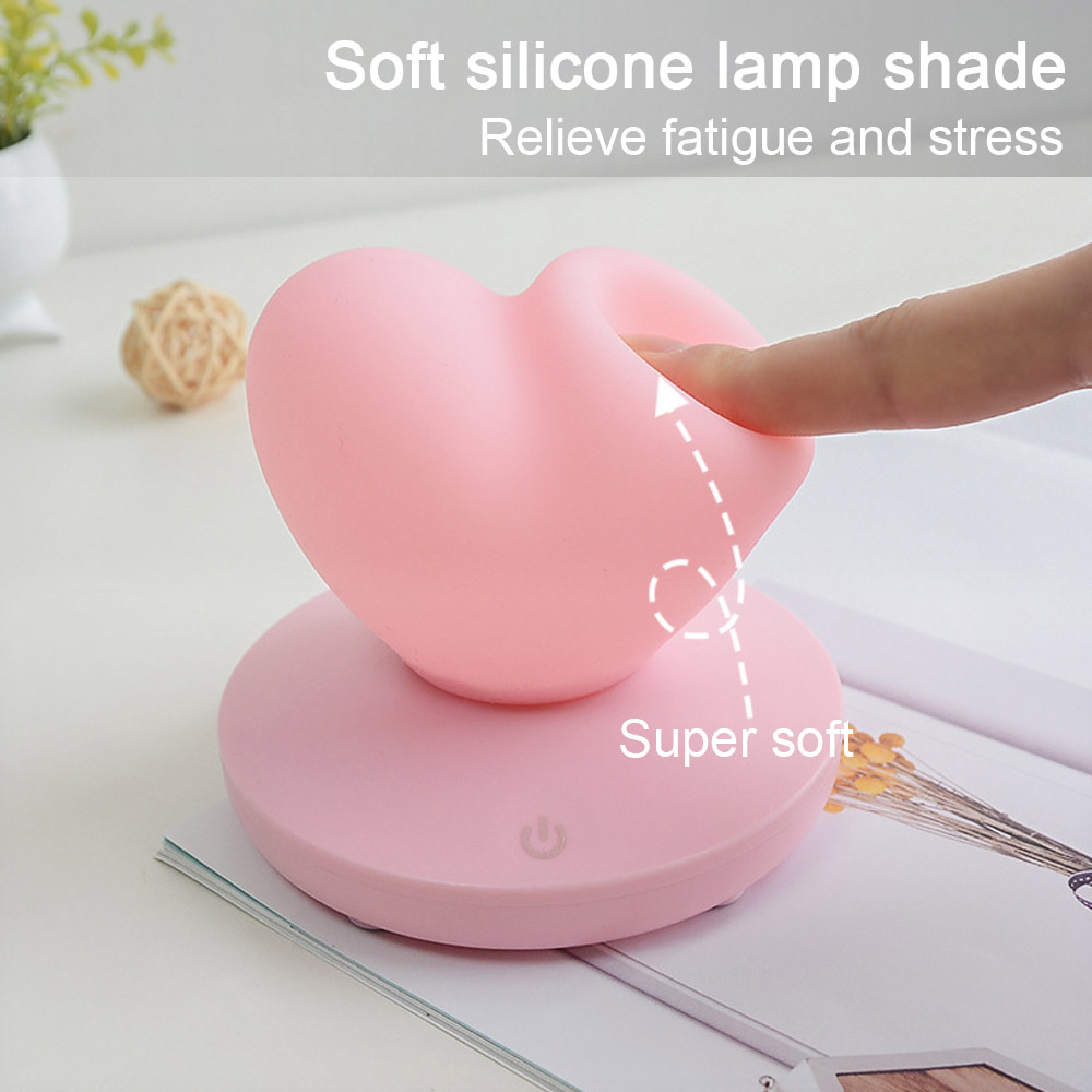 Eine Hand drückt in eine weiche, rosa, herzförmige Silikonlampe auf einem runden Sockel.