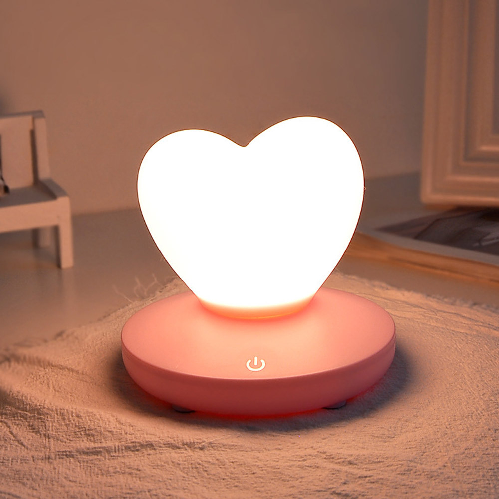 Eine herzförmige Nachtlampe leuchtet auf einem rosa Sockel auf einer weißen Unterlage.