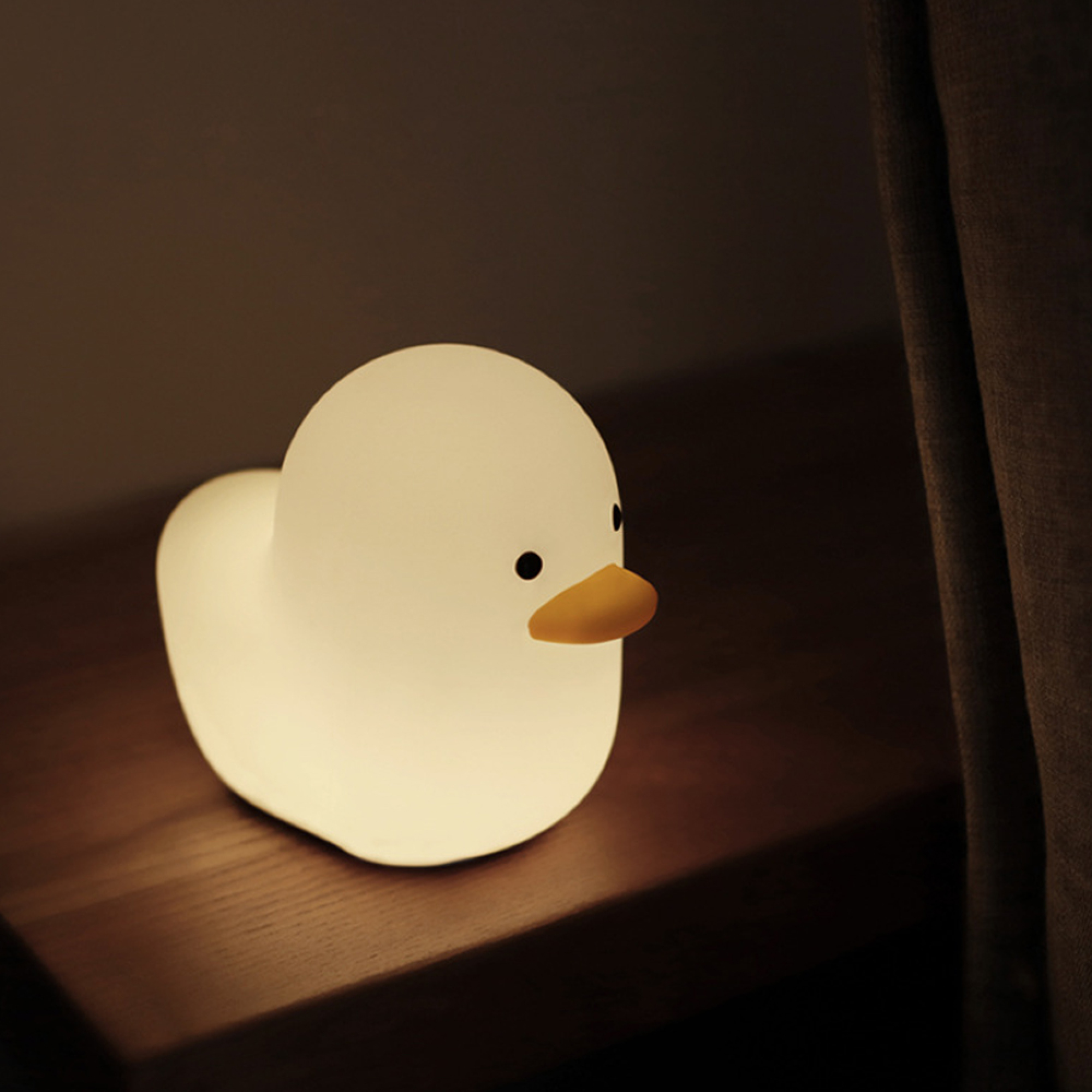 Eine kleine, glühende Entenlampe steht auf einem dunklen Holztisch neben einem Vorhang.