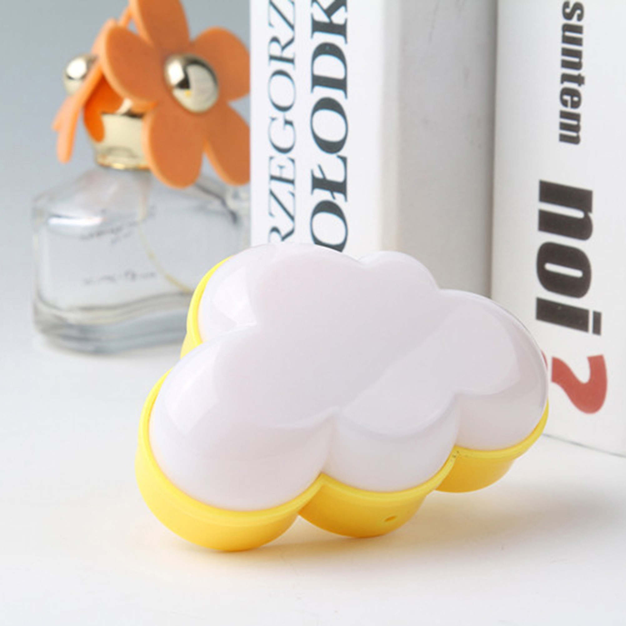 Ein weißes, wolkenförmiges Plastikobjekt mit gelbem Sockel liegt auf weißer Oberfläche, mit Büchern und Parfümflakon.