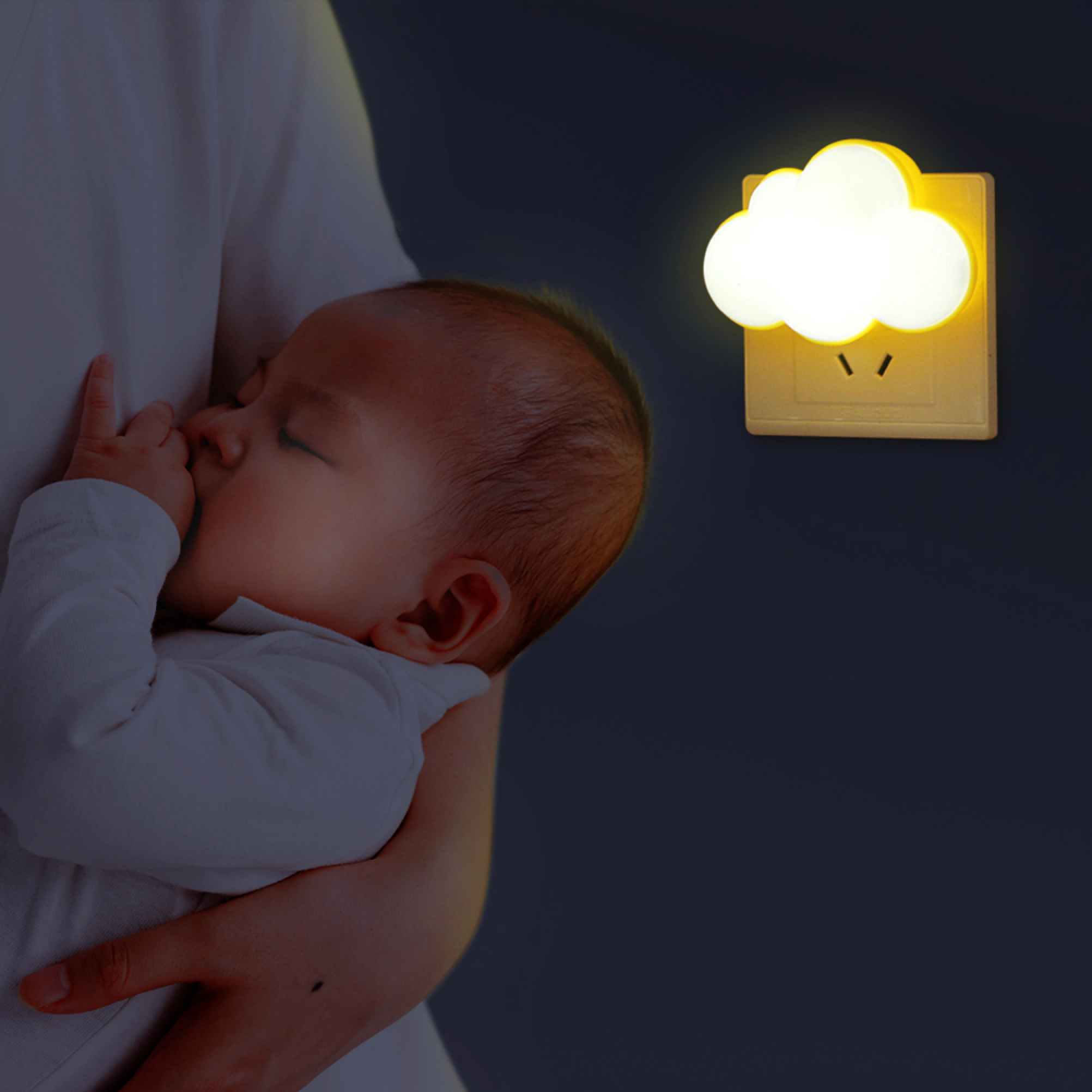 Ein Baby schläft friedlich in den Armen einer Person, während an der dunklen Wand ein gelbes Wolken-Nachtlicht leuchtet.