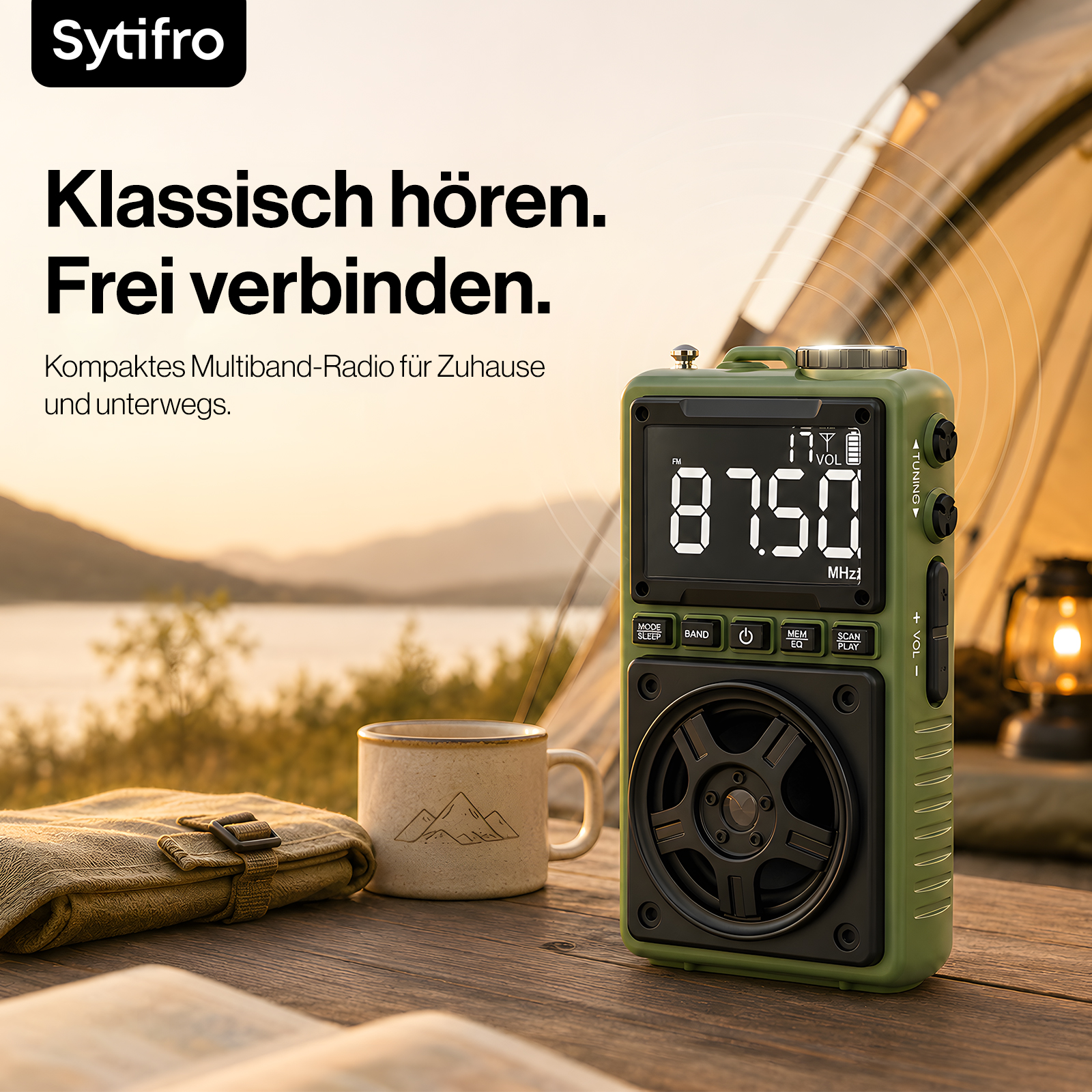 Ein grünes Multiband-Radio, eine Tasse und Stoff auf einem Holztisch draußen vor einem Zelt und See.