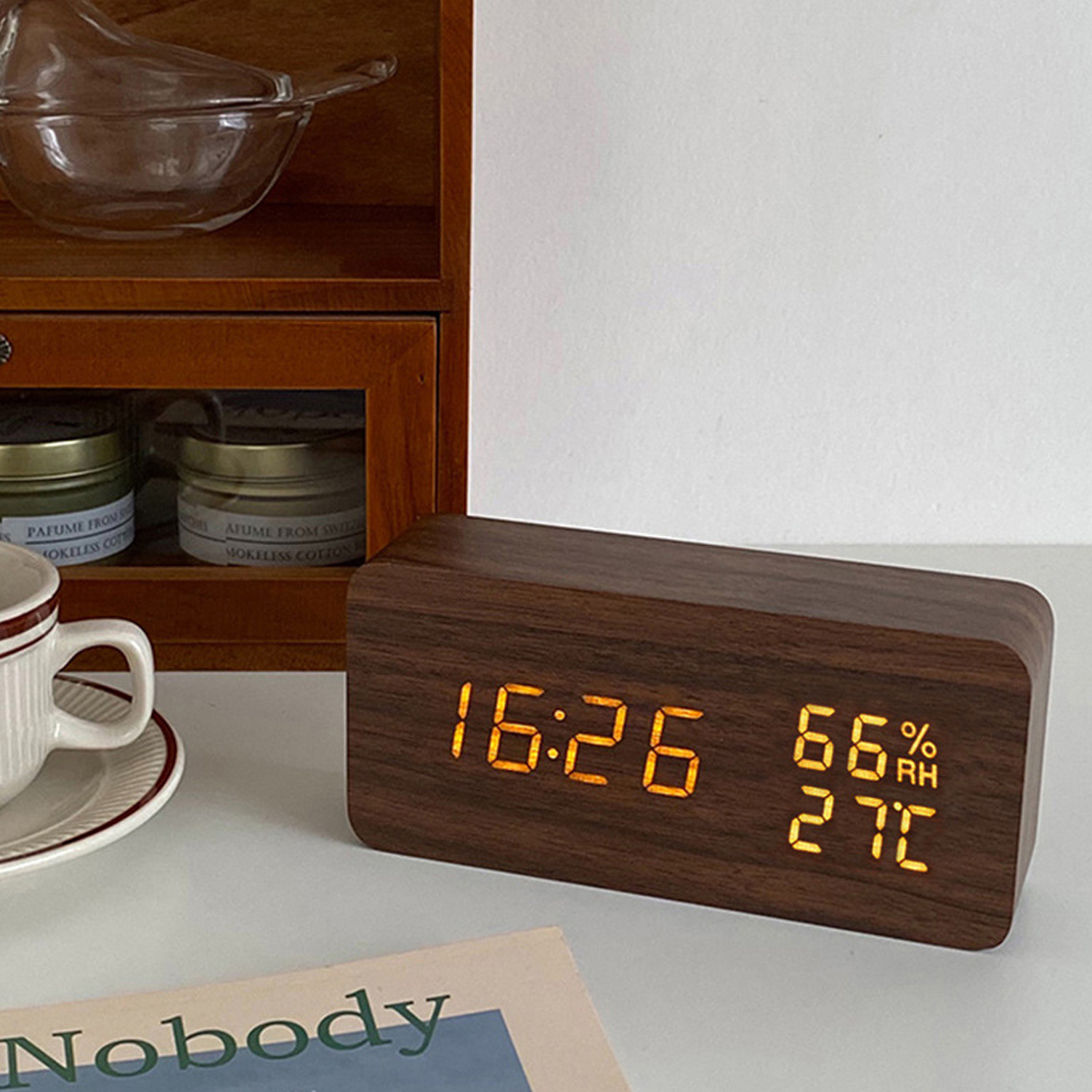 Eine hölzerne Digitaluhr zeigt 16:26, 66% Luftfeuchtigkeit und 27 Grad an, neben einer Tasse und einem Regal.
