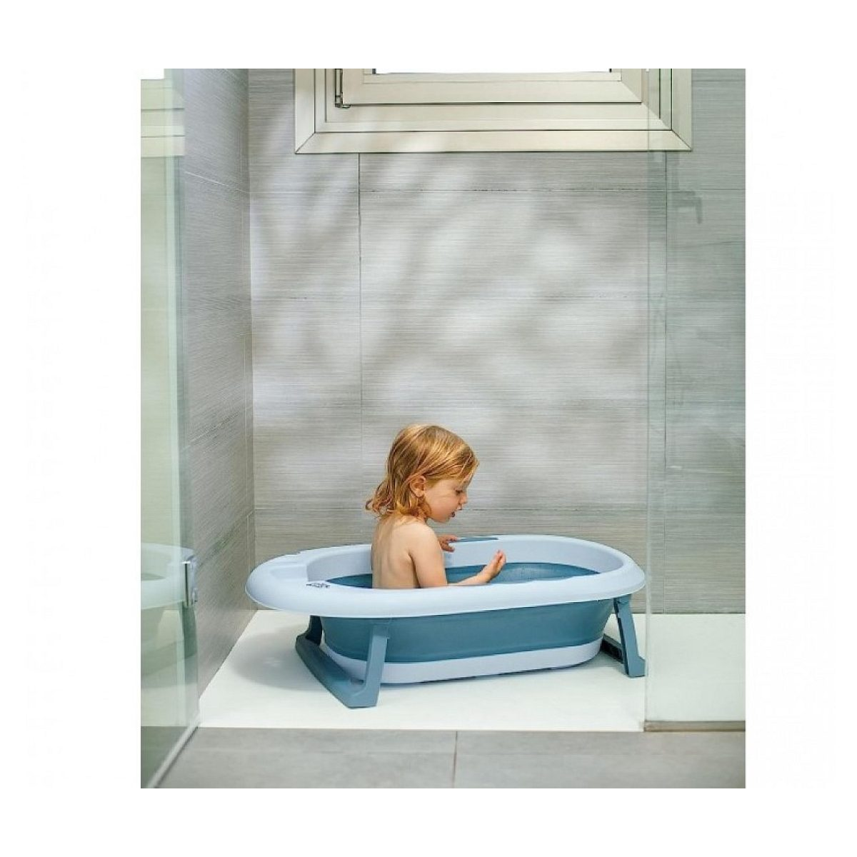 Ein kleiner Knirps mit blonden Haaren sitzt in einer hellblauen und dunkelblauen klappbaren Babybadewanne.