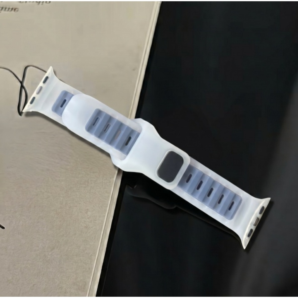 Ein weißes Silikon-Smartwatch-Armband mit grauen Rillen liegt diagonal auf beige-schwarzem Hintergrund.